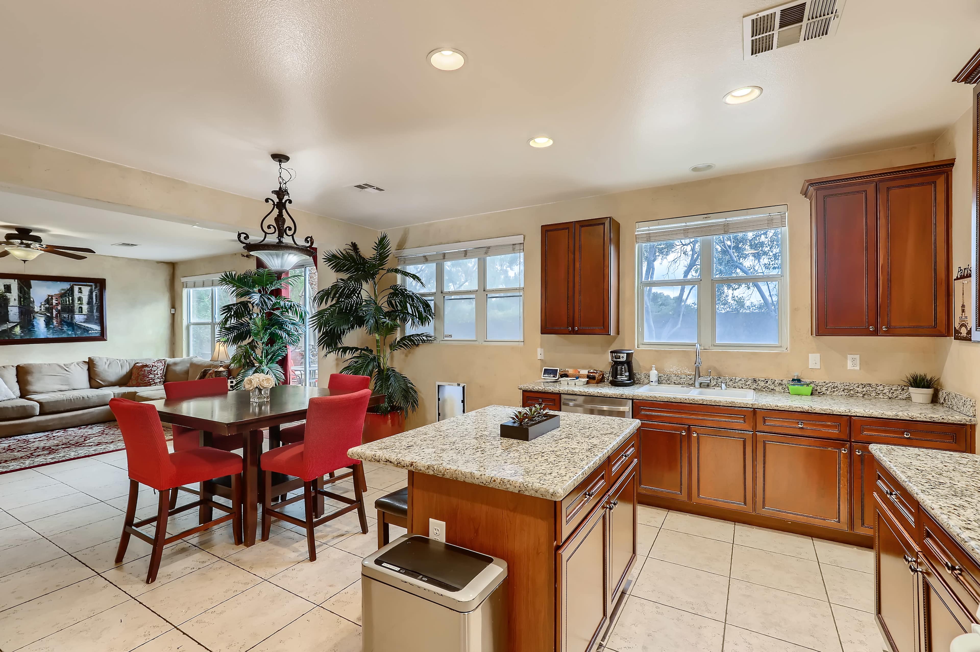 A kitchen with wooden cabinets, a granite countertop, and a dining area featuring a table with red chairs and a living room visible in the background.