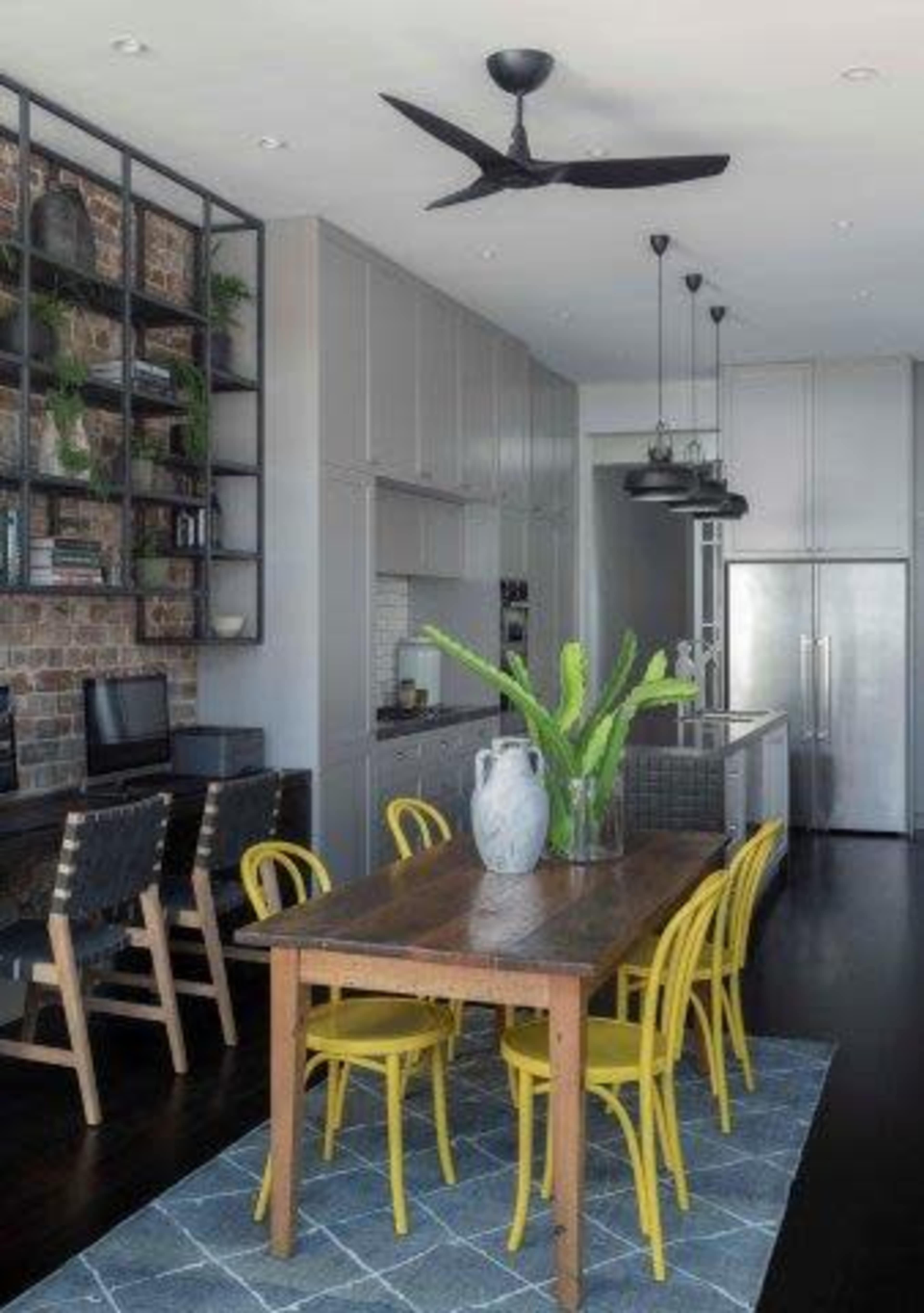 The image shows a modern kitchen and dining area with a wooden table surrounded by yellow chairs, a brick wall feature, and sleek cabinetry.