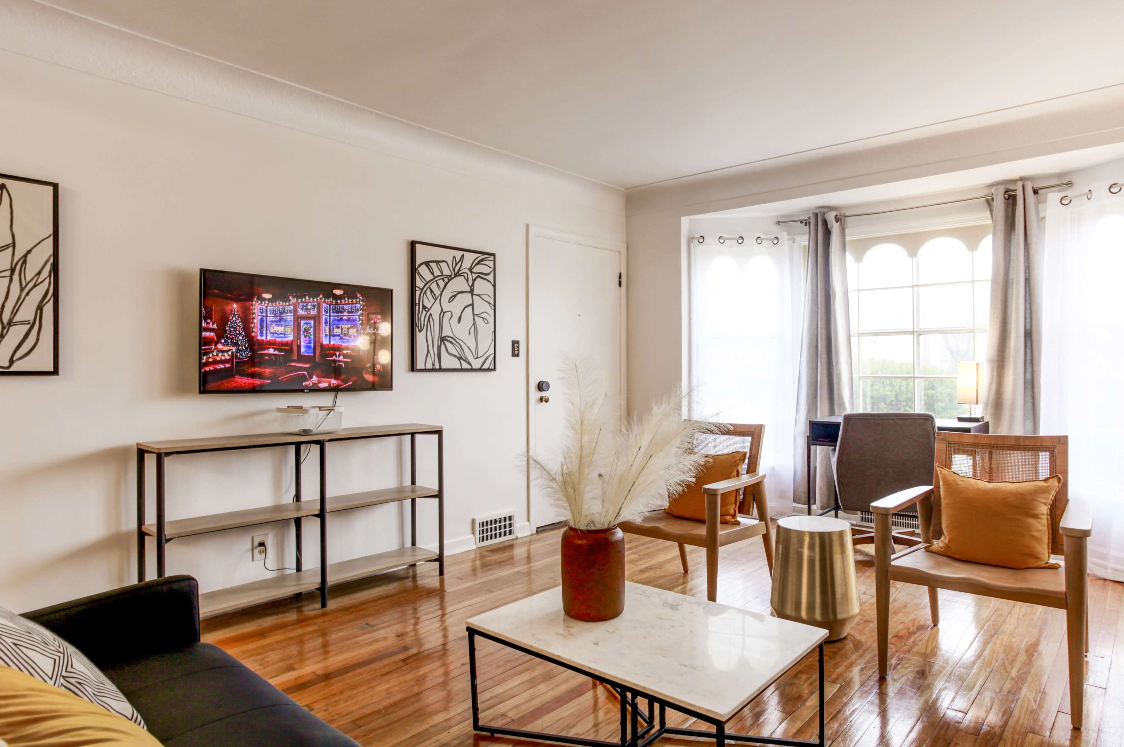 A well-lit living room features a television mounted on the wall, a coffee table with a decorative vase, and seating arranged around a window.