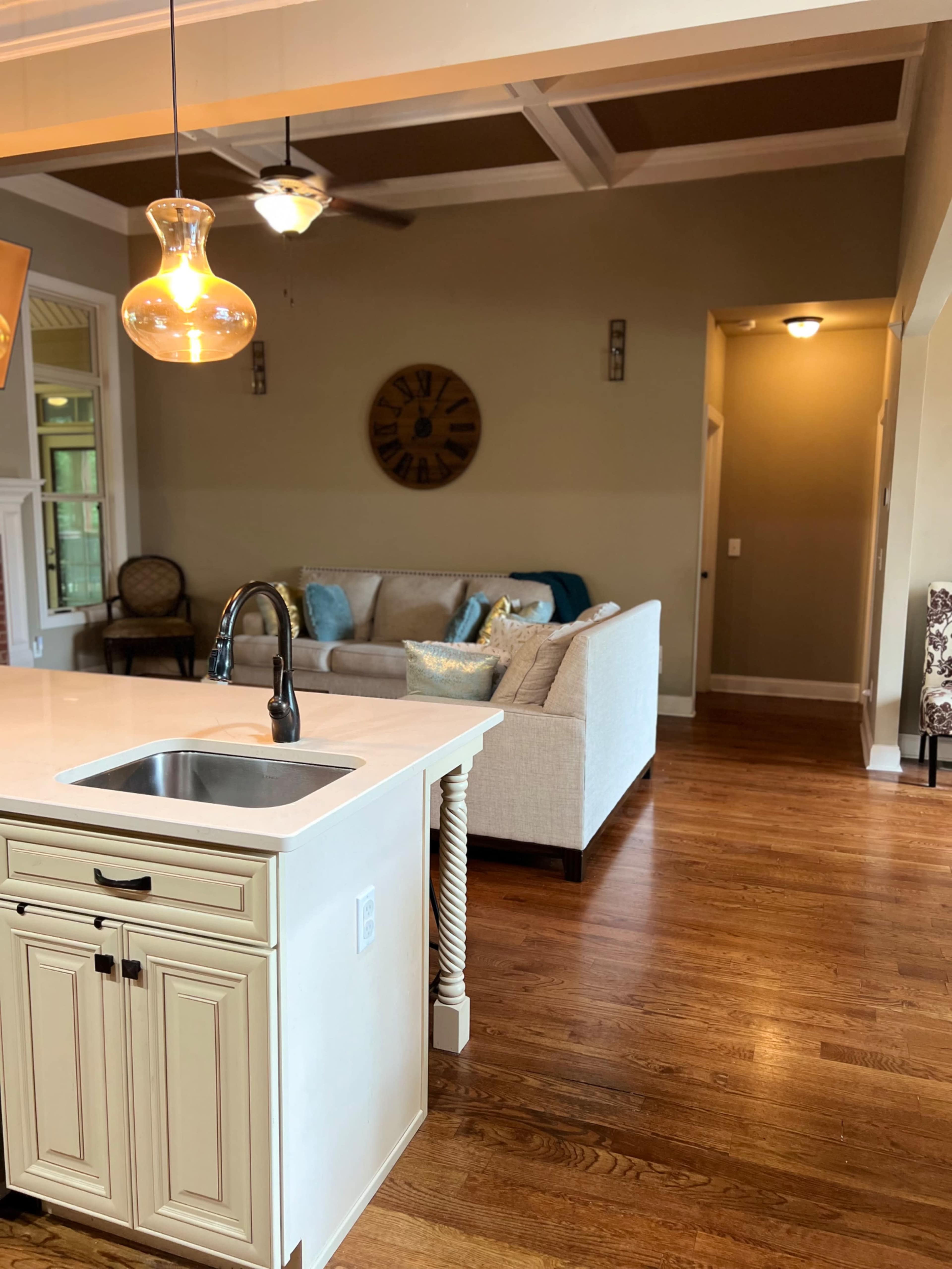 The image shows a modern kitchen with a white island sink, wooden flooring, and a cozy living area featuring a light-colored sofa and decorative pillows in the background.