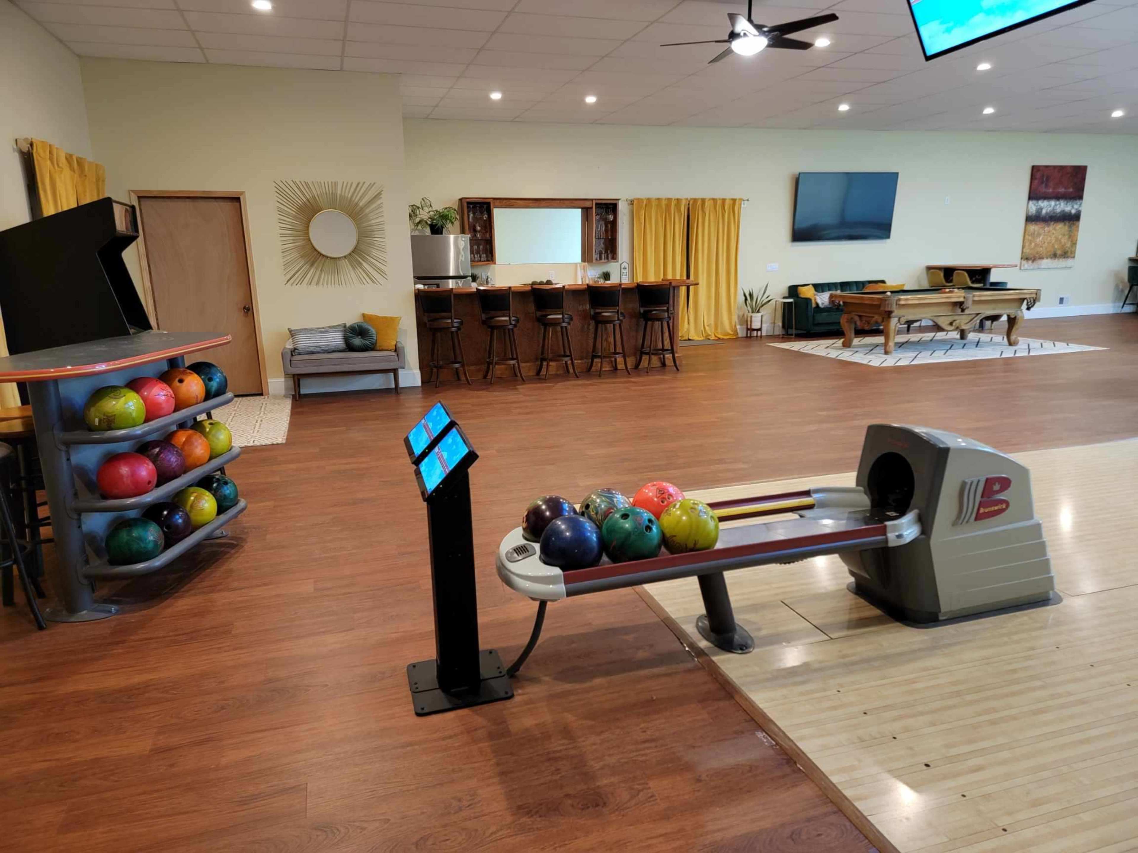 A bowling area featuring a bowling lane with a ball return system and a rack of colorful bowling balls in a spacious room with a bar and seating.