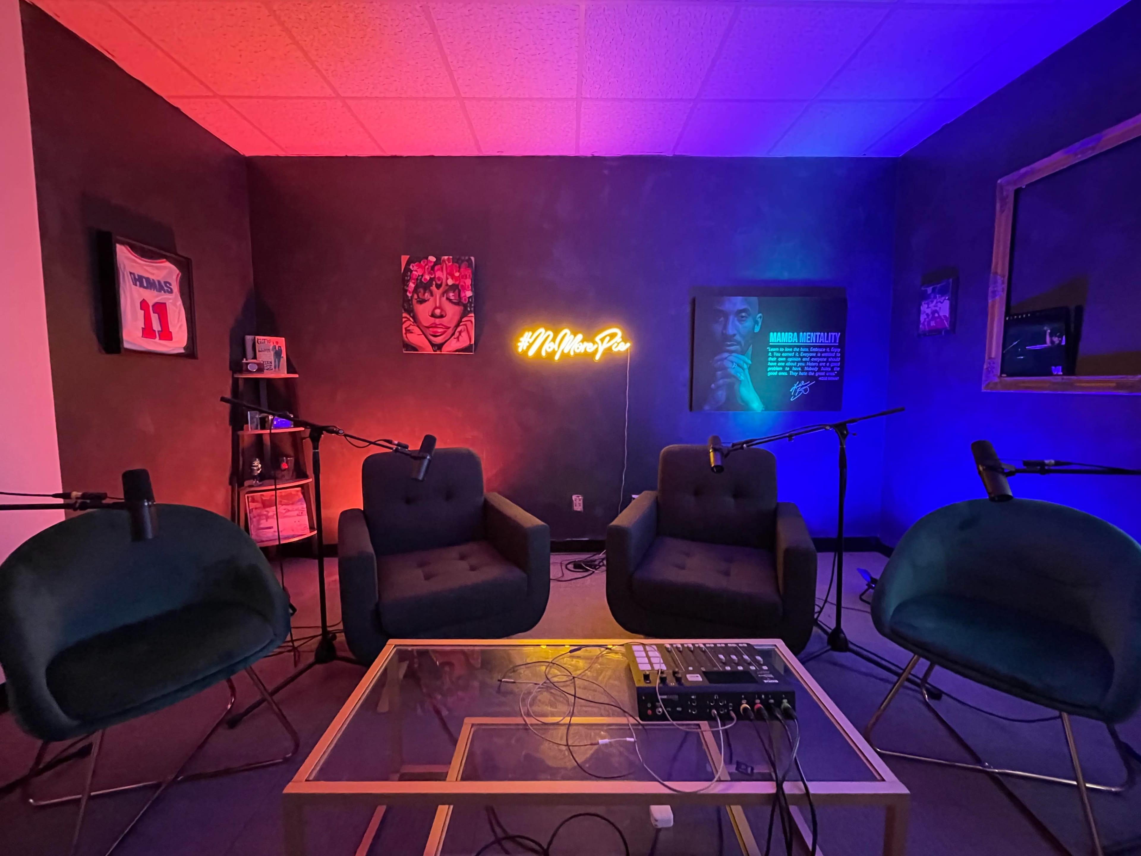 A small, modern studio space with two green chairs, a glass table, and various microphones set up against a backdrop featuring colorful lighting and framed artwork.