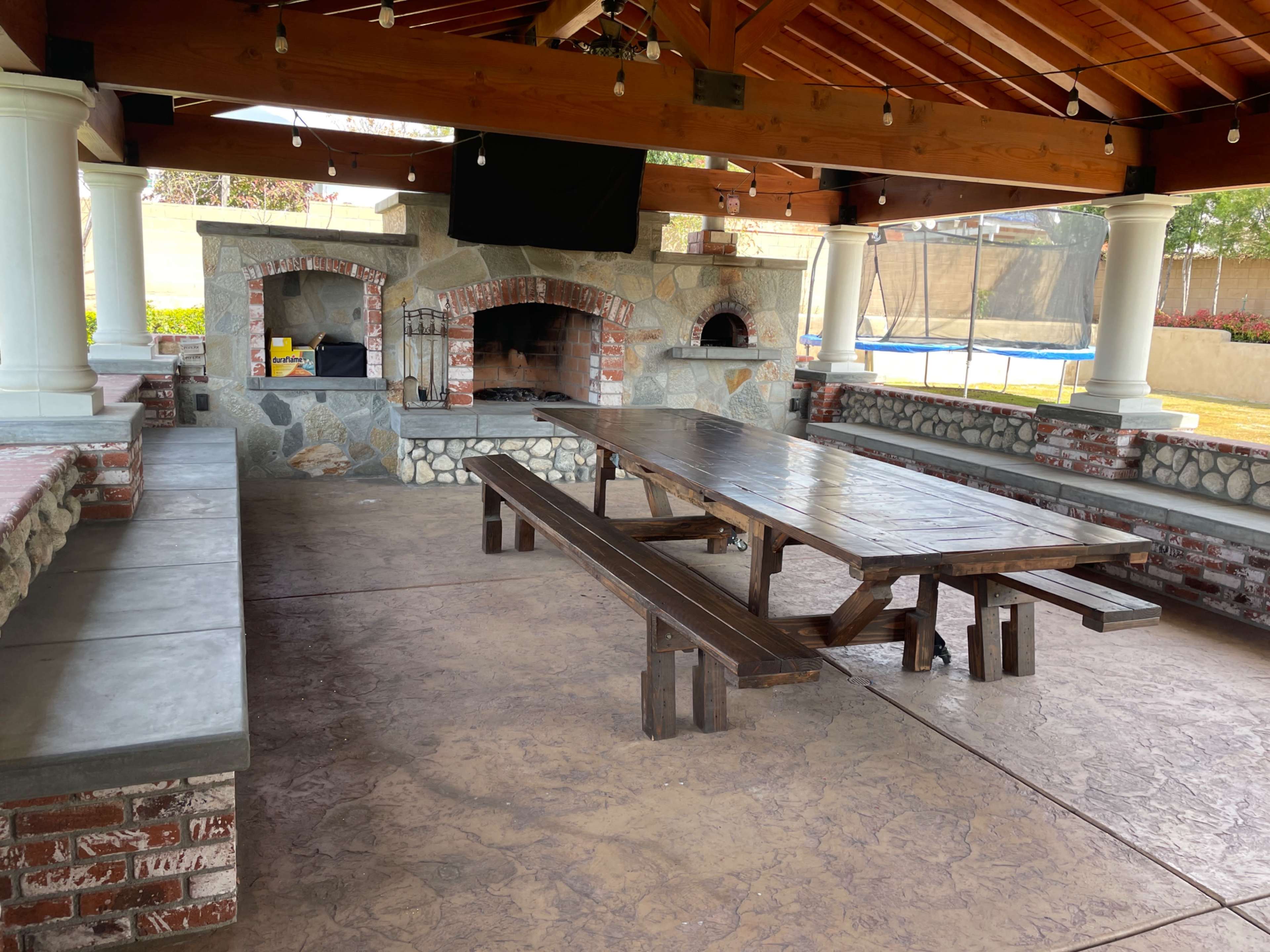 A spacious outdoor patio with a large wooden table, stone fireplace, and a background featuring a trampoline.
