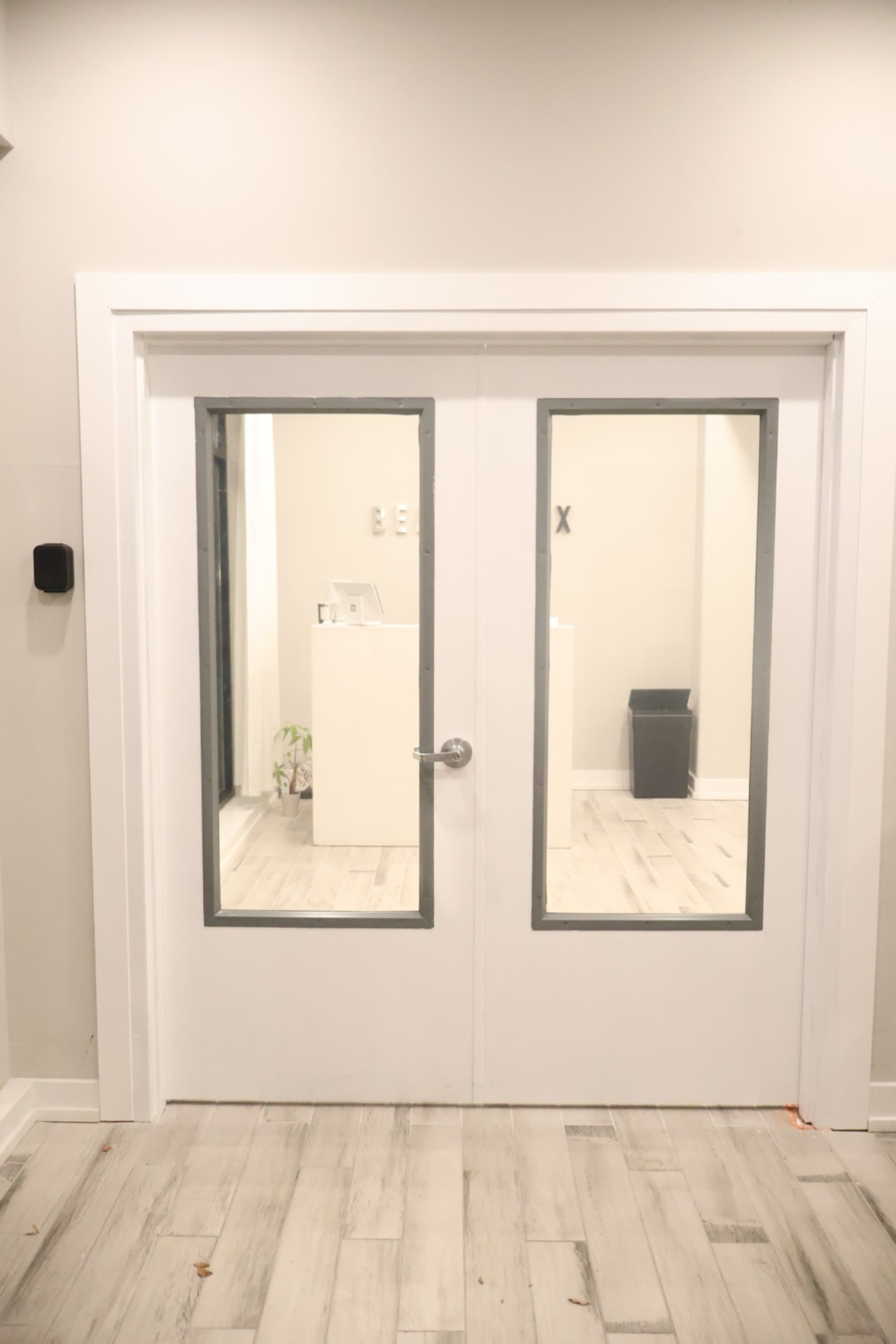 The image shows a double glass door with a silver handle, leading into a brightly lit room with light-colored walls and flooring.