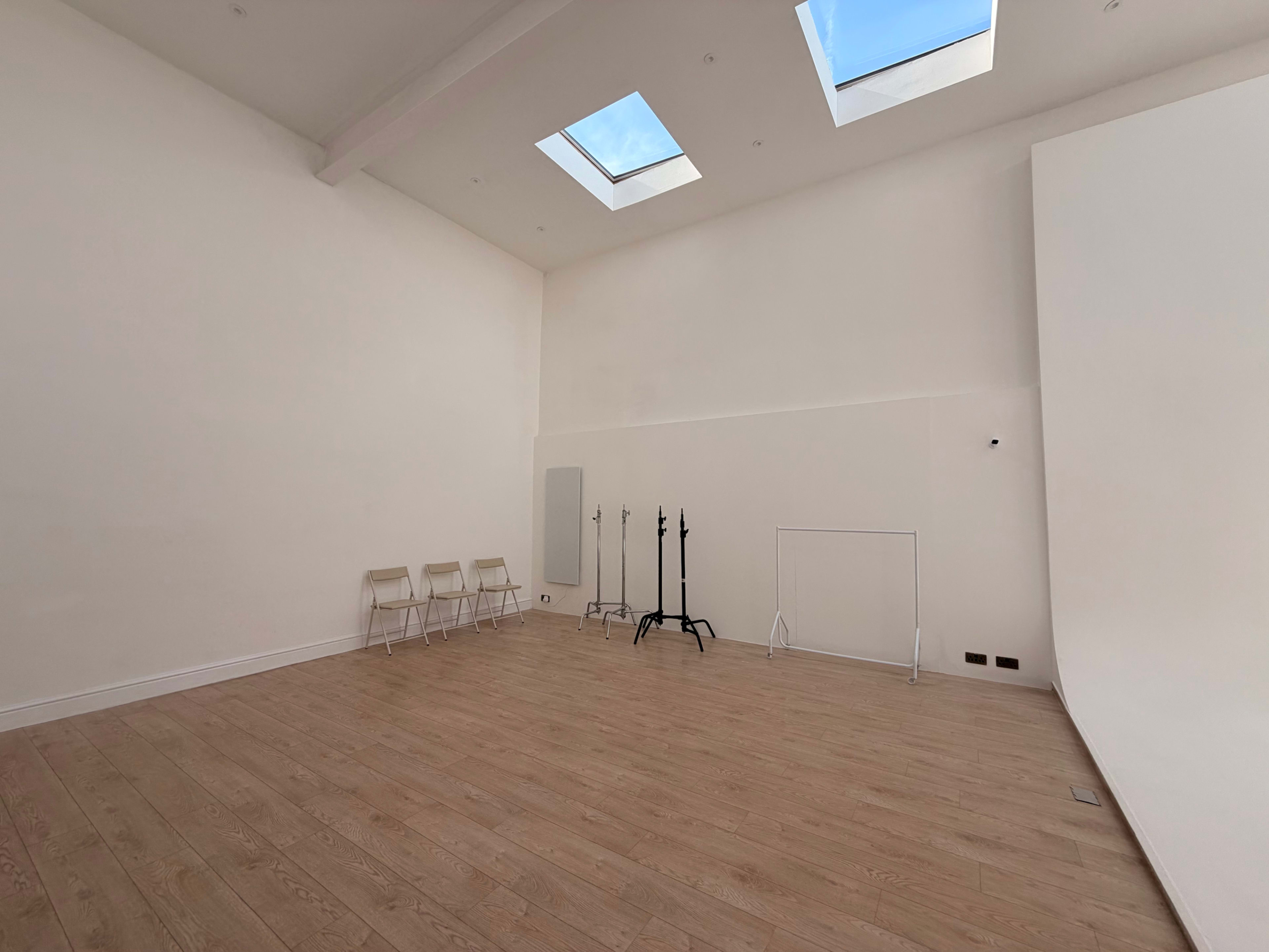 The image shows a spacious, empty room with wooden flooring, white walls, skylights, and several folding chairs along one side.