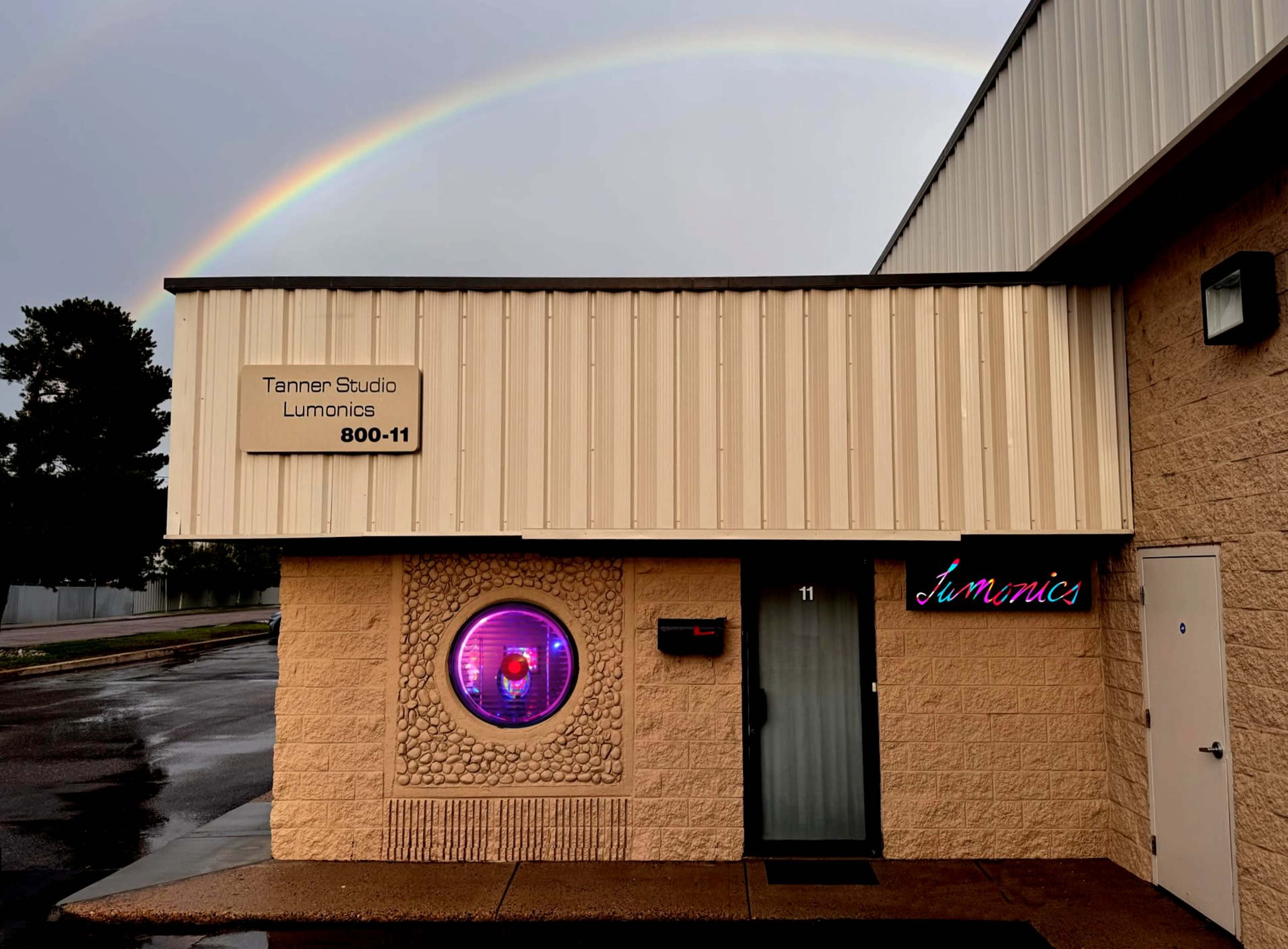 A building with a sign reading "Tannen Studio Lumonics" and a circular window displays an illuminated design, under an arching rainbow.
