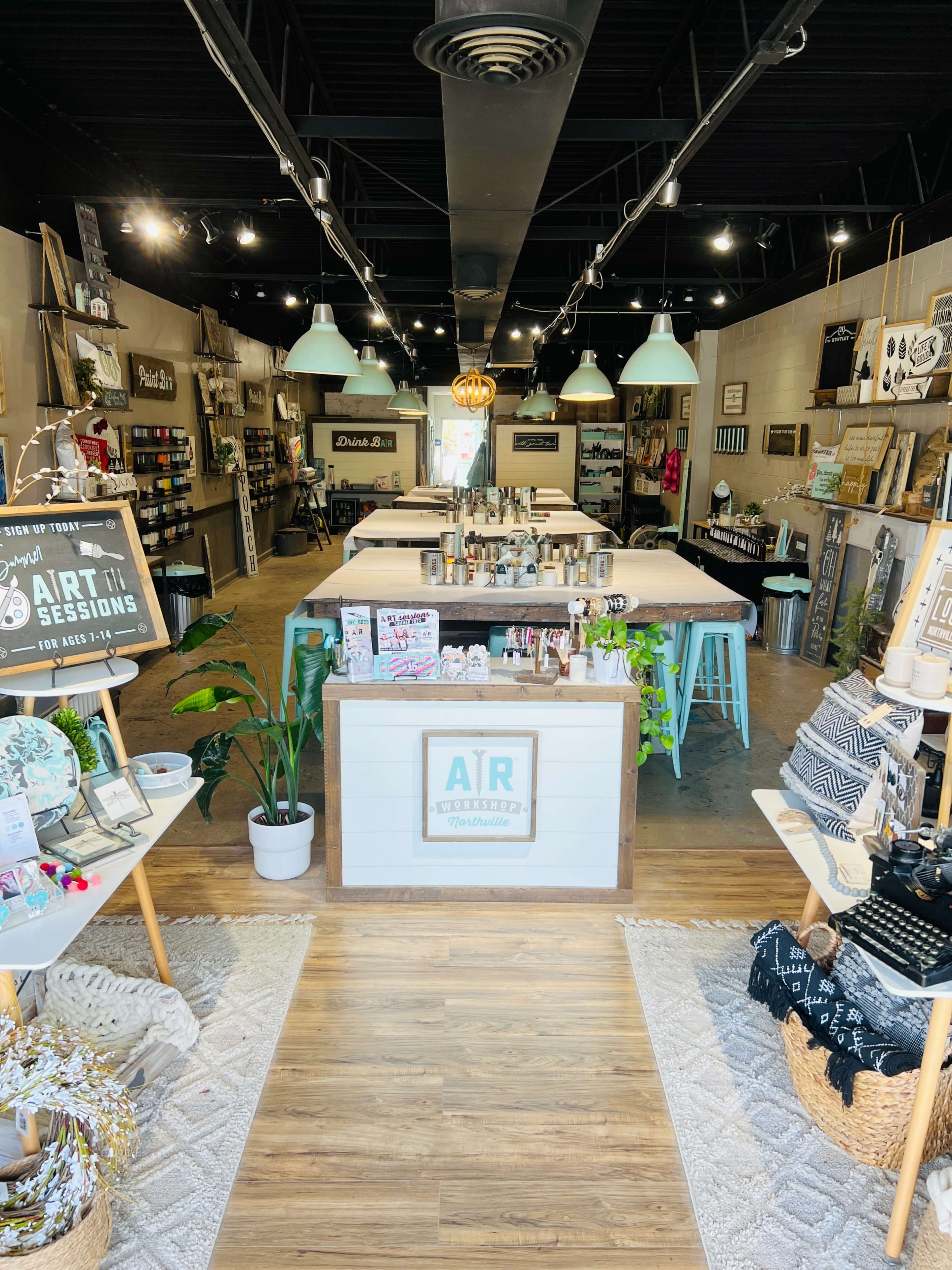 A brightly lit shop interior featuring a central display table with art supplies, surrounded by various shelves and products, and a welcoming entrance.