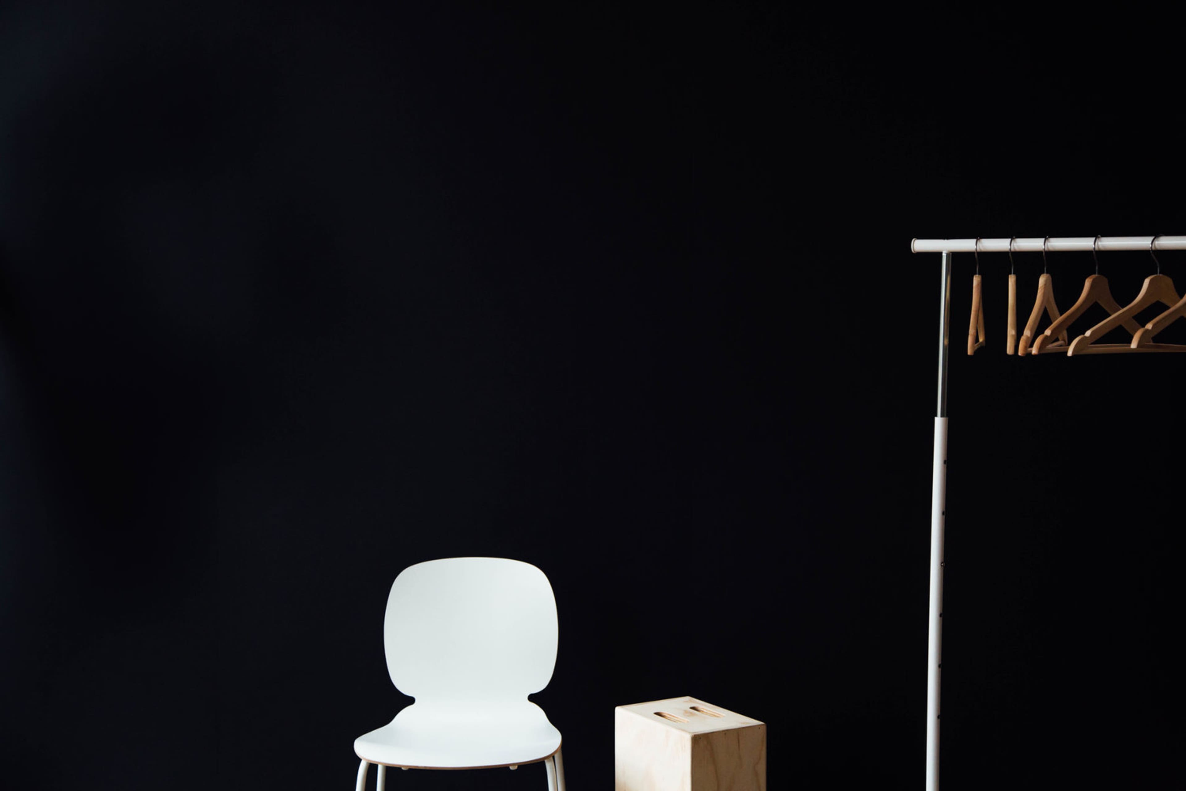 A white chair is positioned next to a wooden box and a clothing rack with hangers against a black wall.