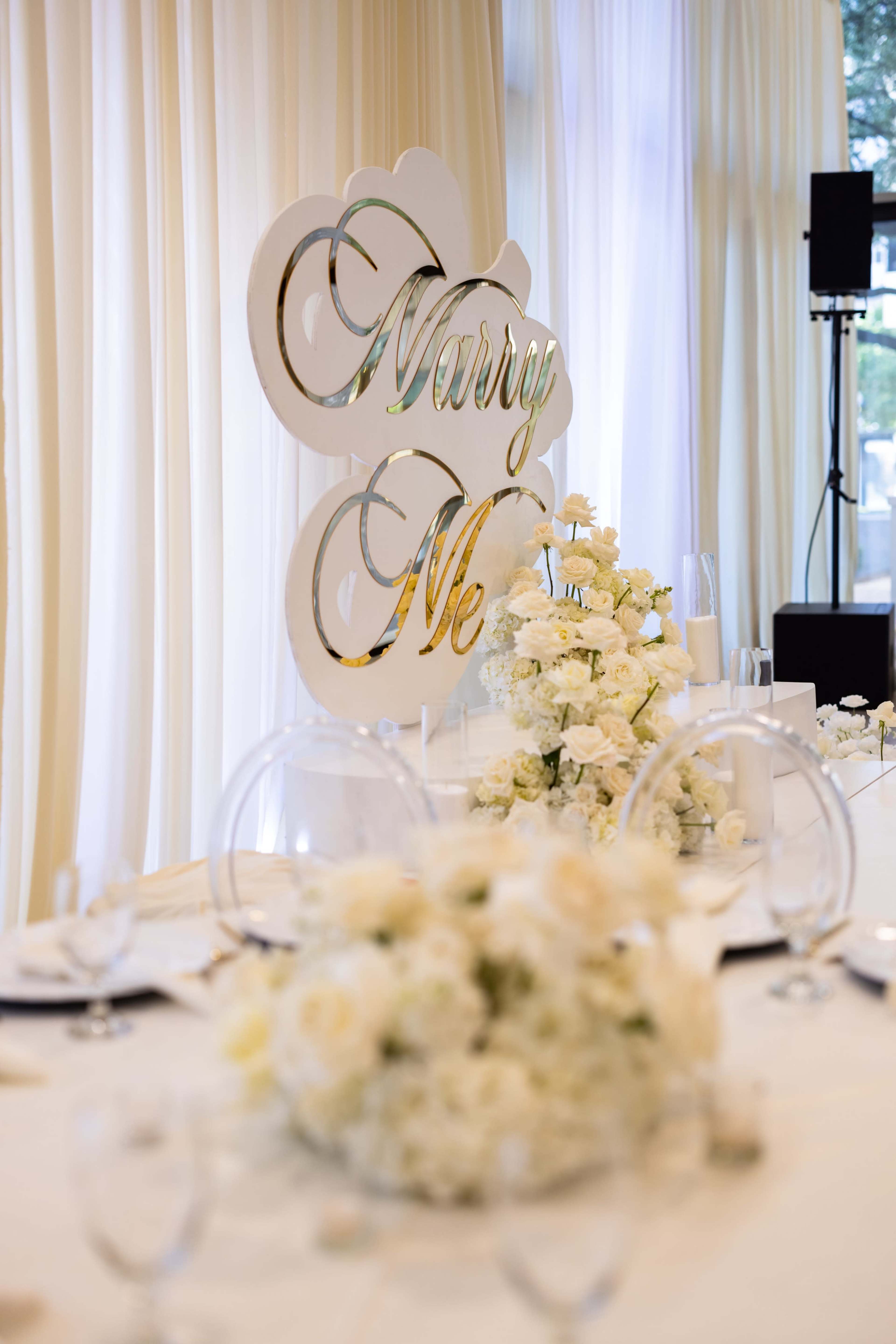 A table decorated with white flowers features a decorative sign that reads "Marry Me" in a prominent display.