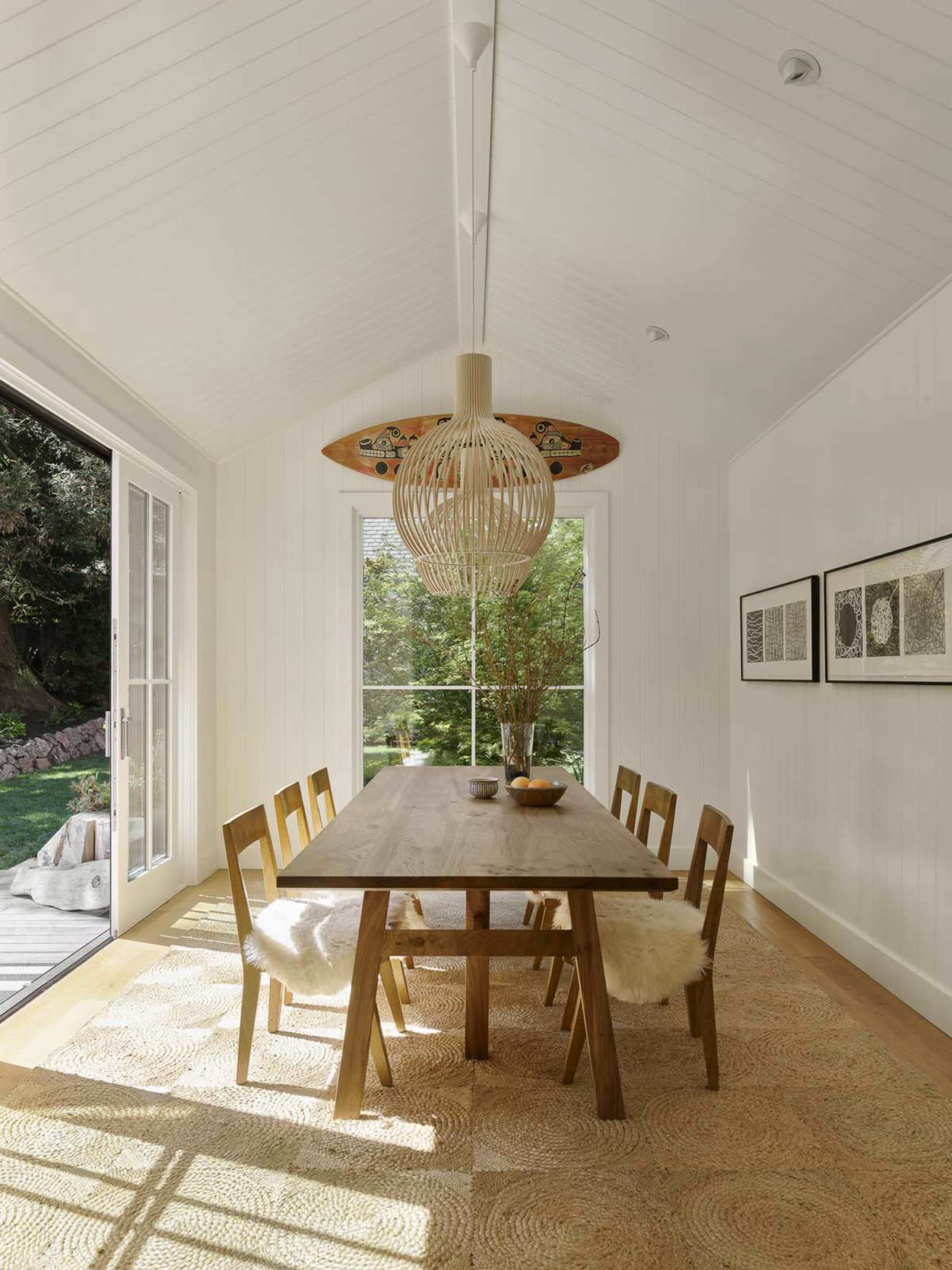 A bright dining area features a wooden table surrounded by six chairs, illuminated by a large woven pendant light, with windows that look out onto greenery.