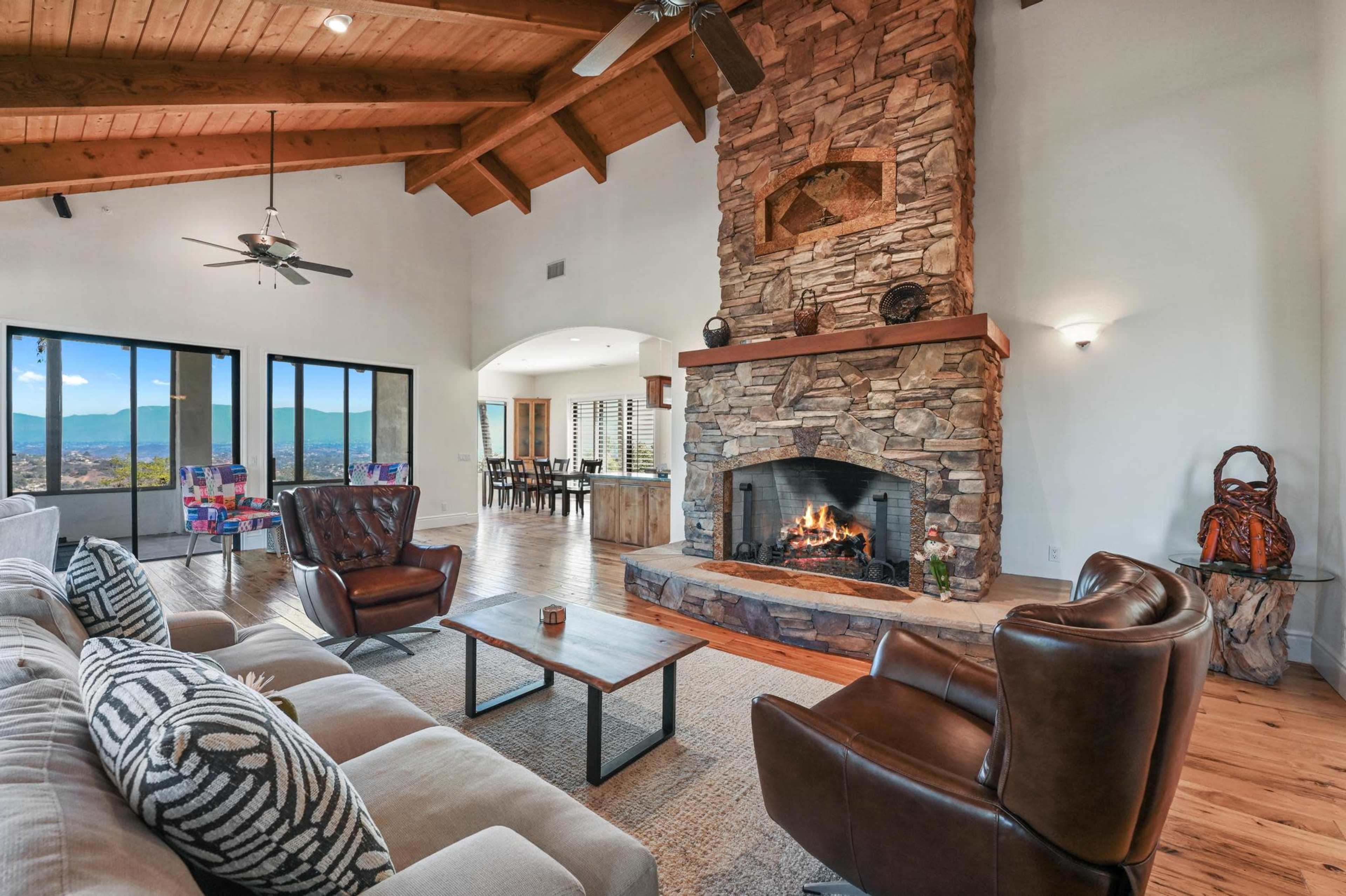 A spacious living room features a stone fireplace, wooden beams, large windows with mountain views, and comfortable seating arrangements.