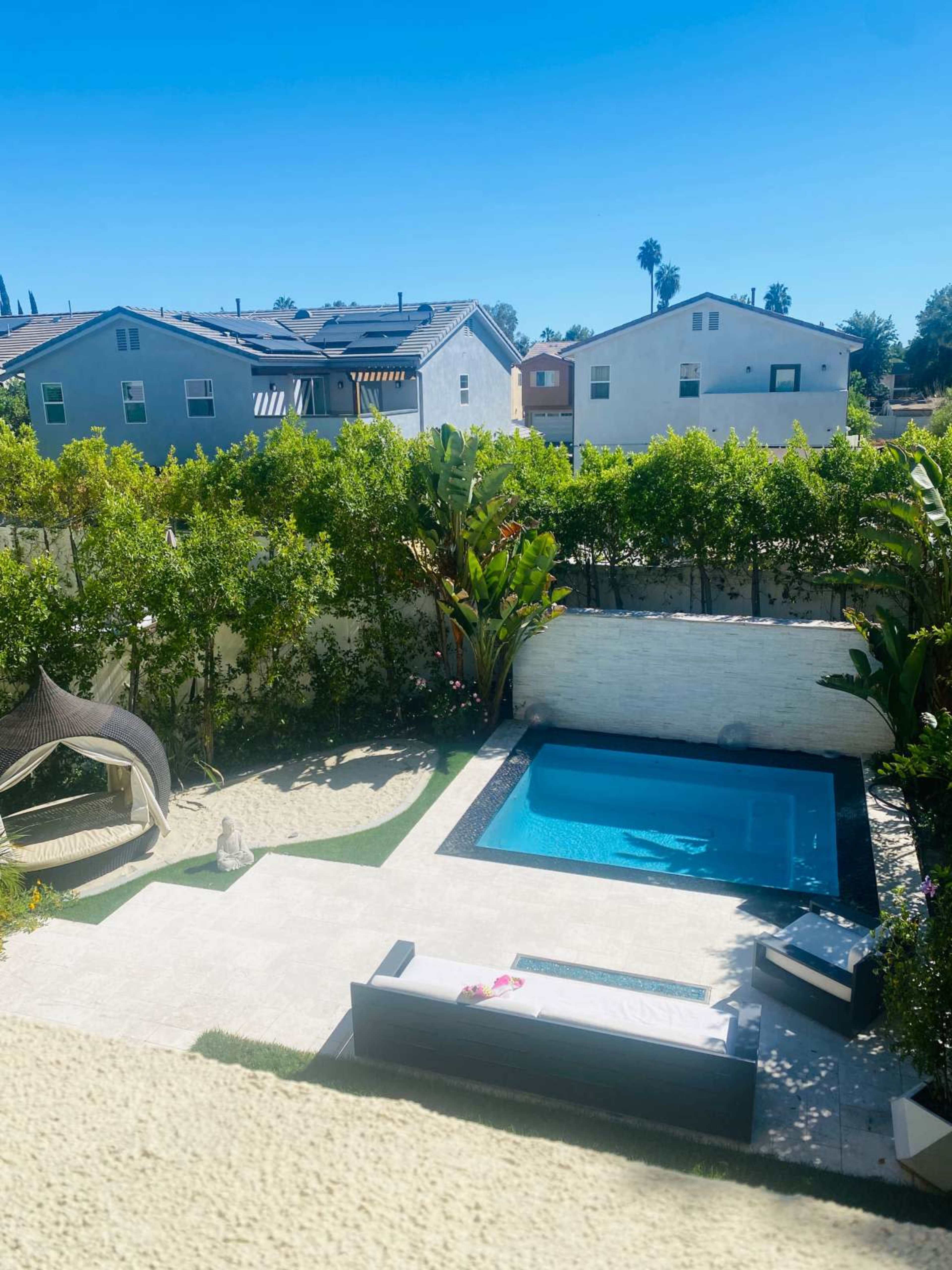 A backyard with a swimming pool, a lounging area, and tropical plants surrounded by a fence.
