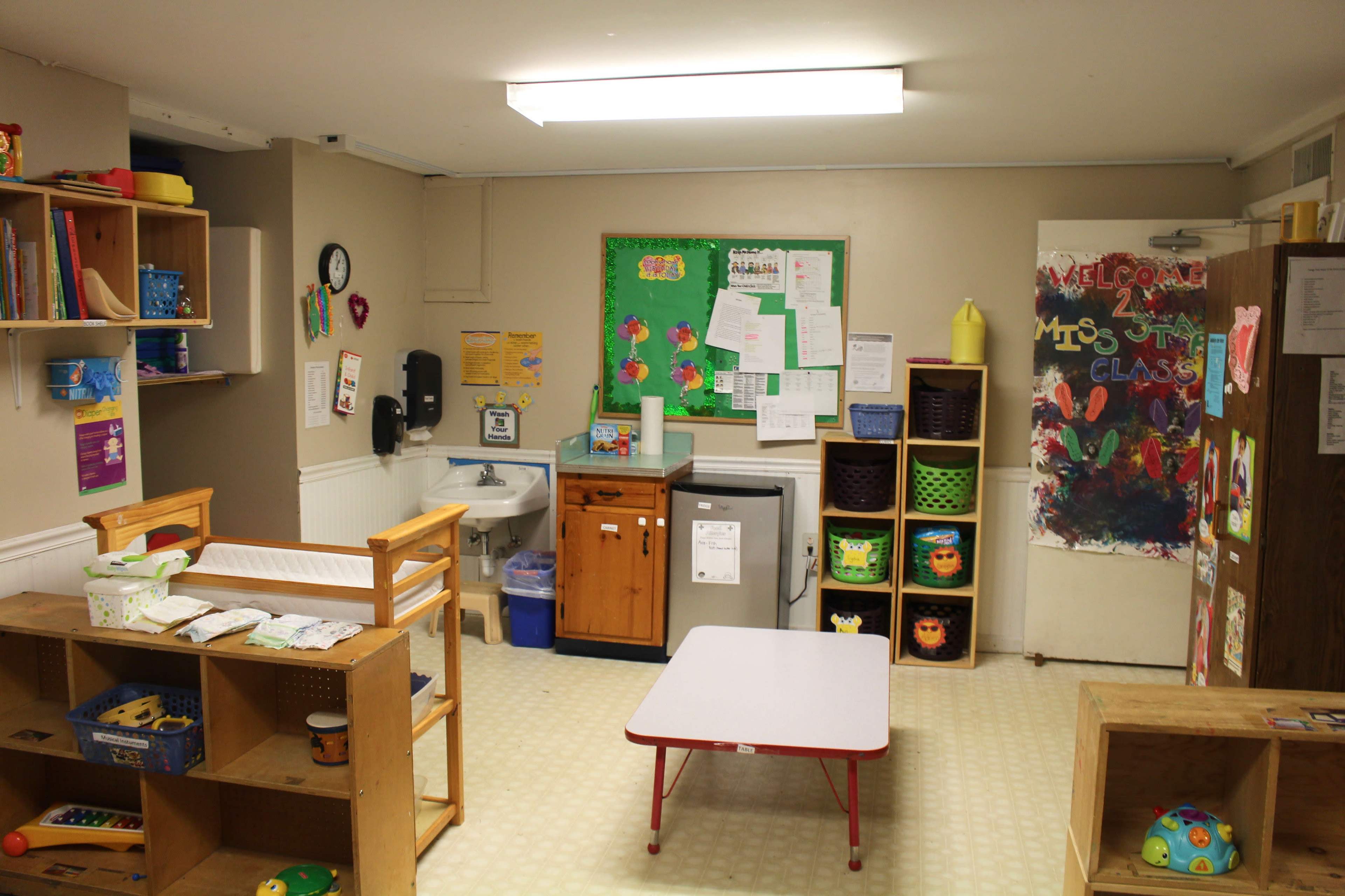 A brightly lit classroom with wooden furniture, a sink, storage cabinets, and colorful organizational bins.