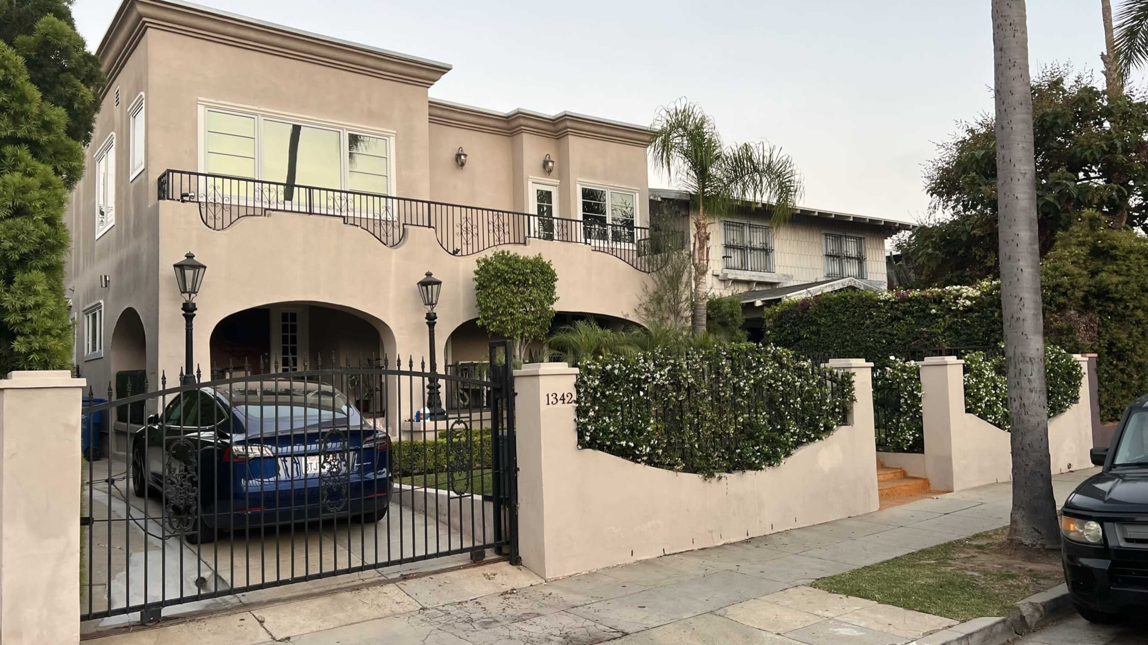 The image shows a two-story beige house with a gated driveway, a manicured front garden, and palm trees, located on a suburban street.