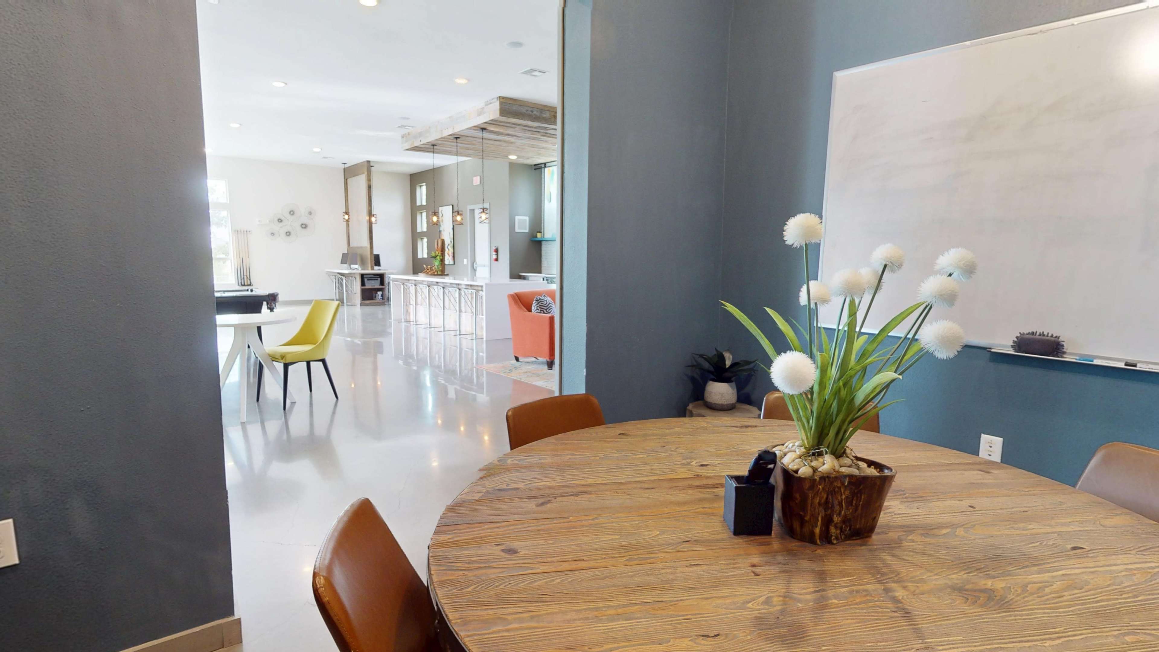 A wooden round table with a plant centerpiece is positioned near a wall, leading into a bright room with a kitchen and dining area visible in the background.