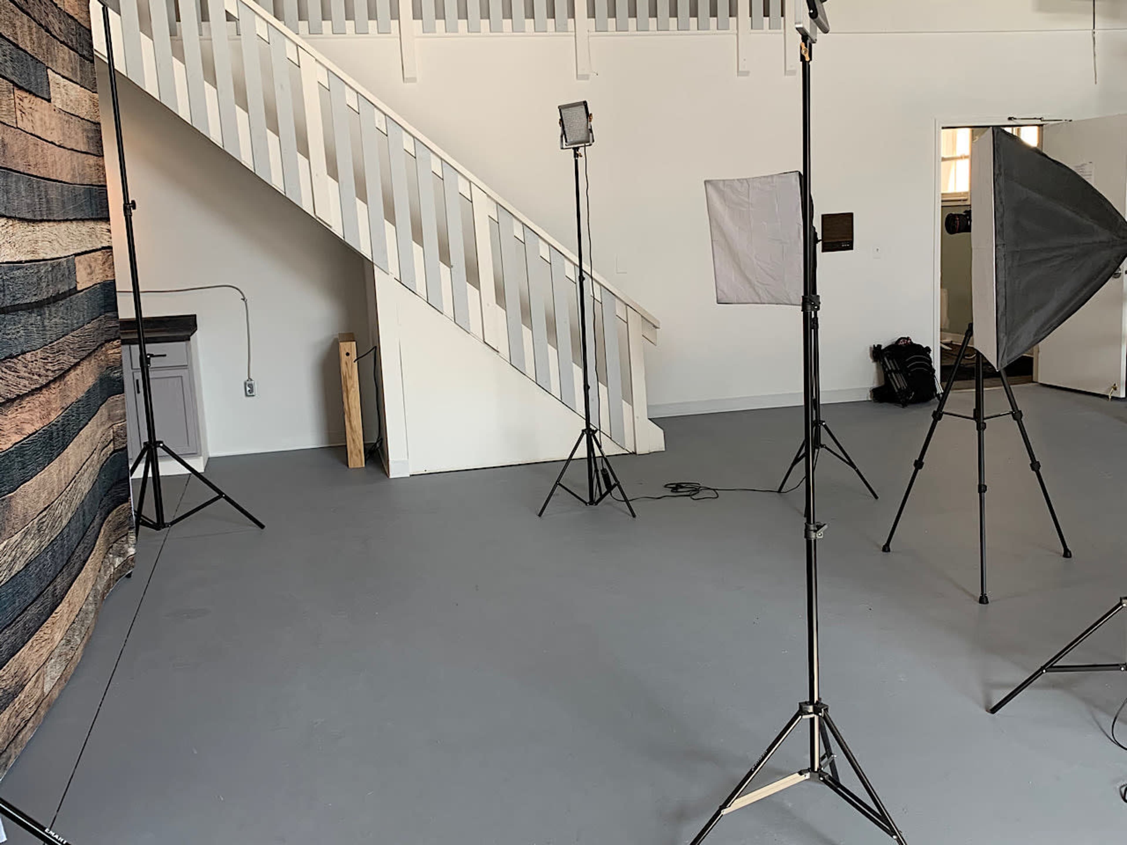 A photography studio setup with several light stands, soft boxes, and a wooden backdrop, alongside a staircase and a white wall.