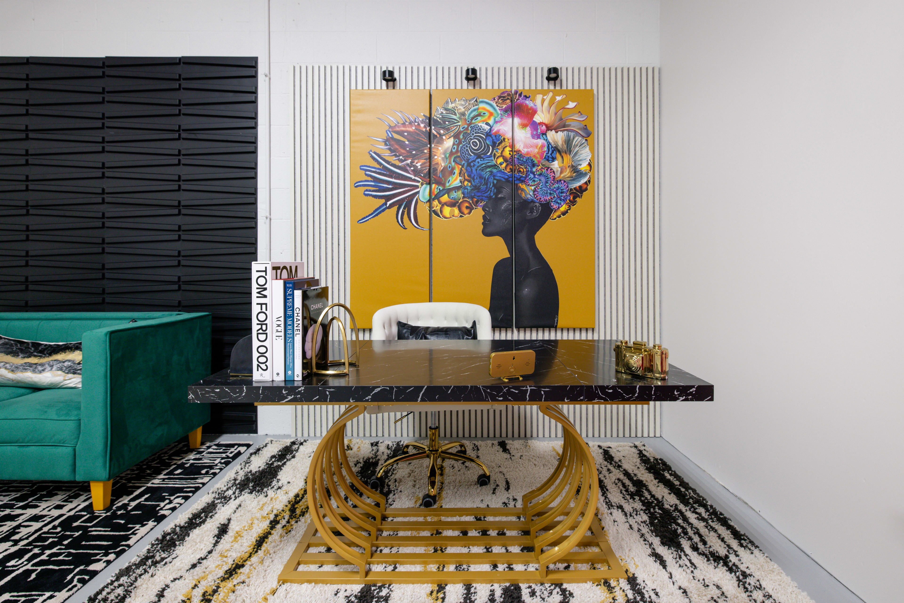 A modern office space featuring a black marble desk with a gold base, a colorful artwork of a woman with an elaborate headdress on the wall, and a green velvet sofa in the background.