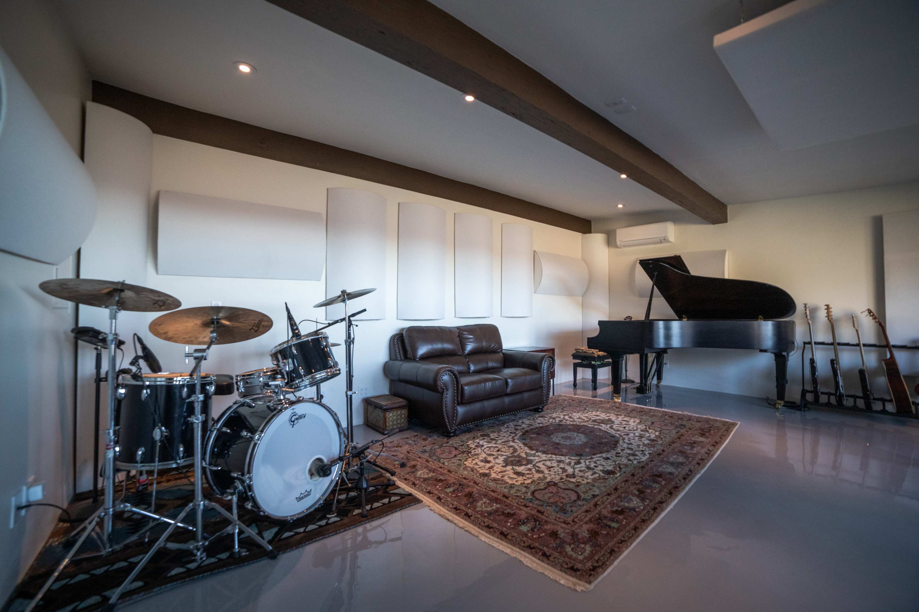 A spacious music studio features a drum set, a grand piano, a leather couch, and several guitars against the wall.