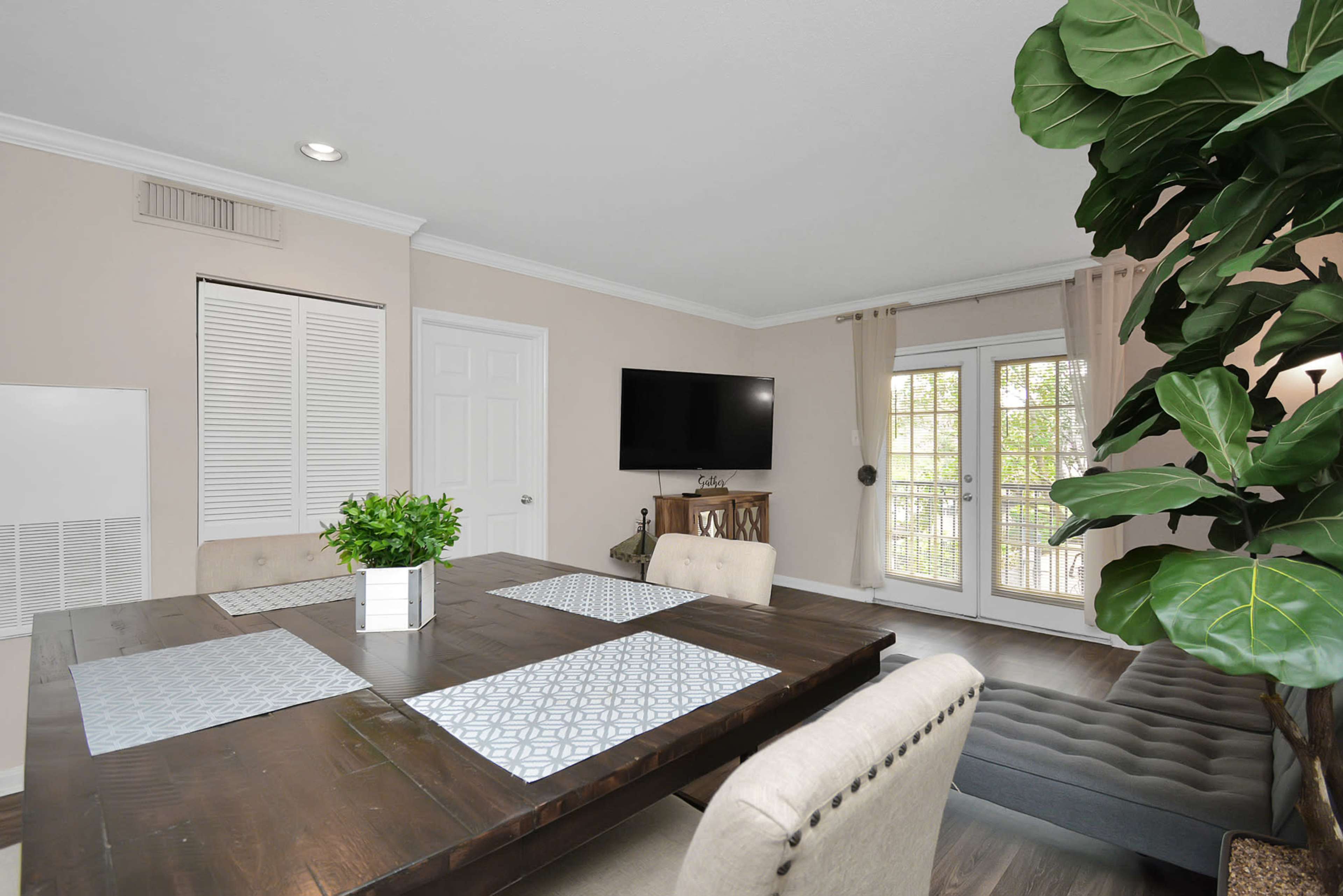 A dining area featuring a wooden table with a patterned table runner, beige chairs, and a large potted plant next to a TV mounted on the wall and French doors leading outside.