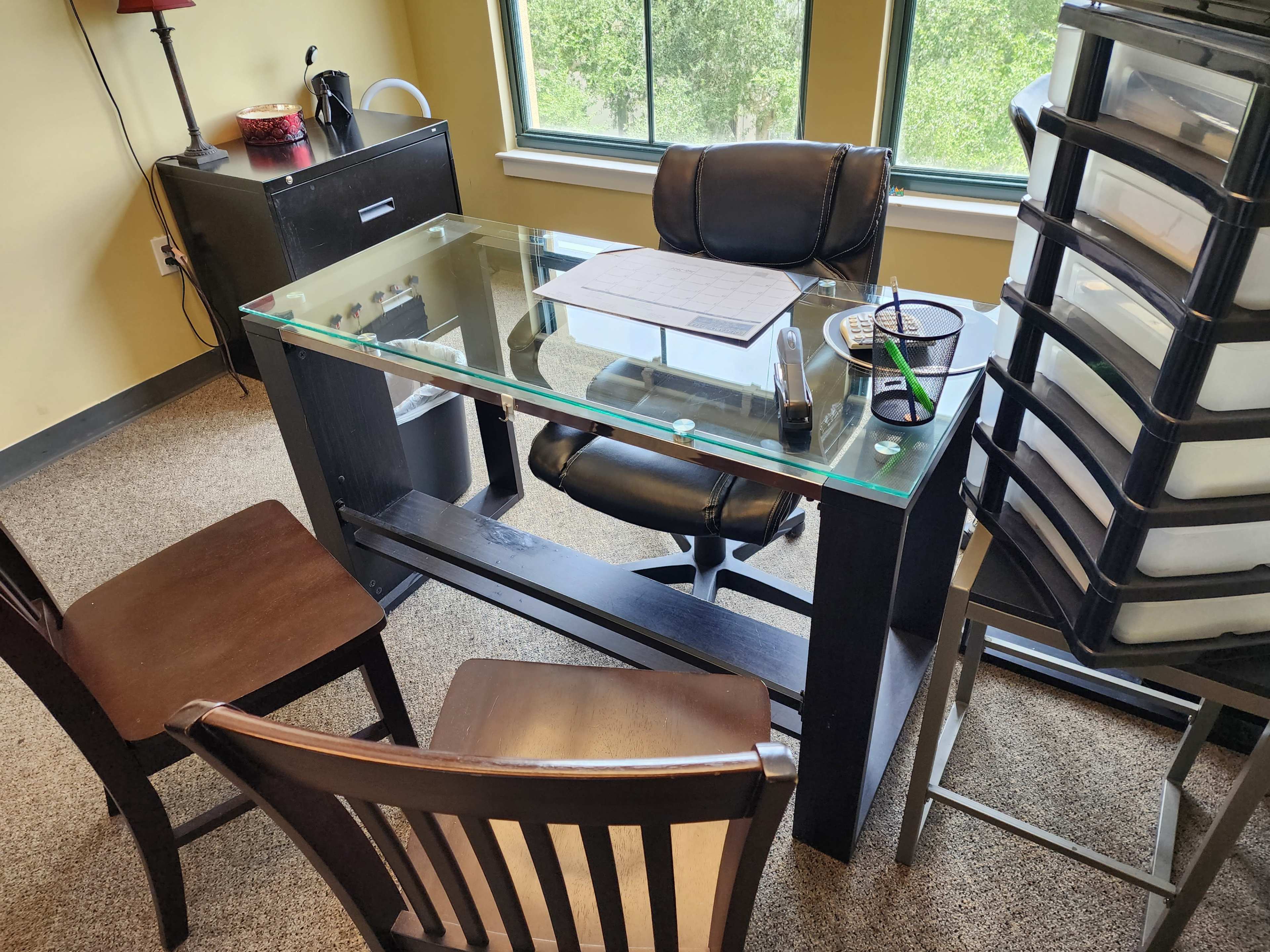 An office scene features a glass-top desk with a chair and two wooden chairs arranged around it, along with storage containers and a window revealing greenery outside.