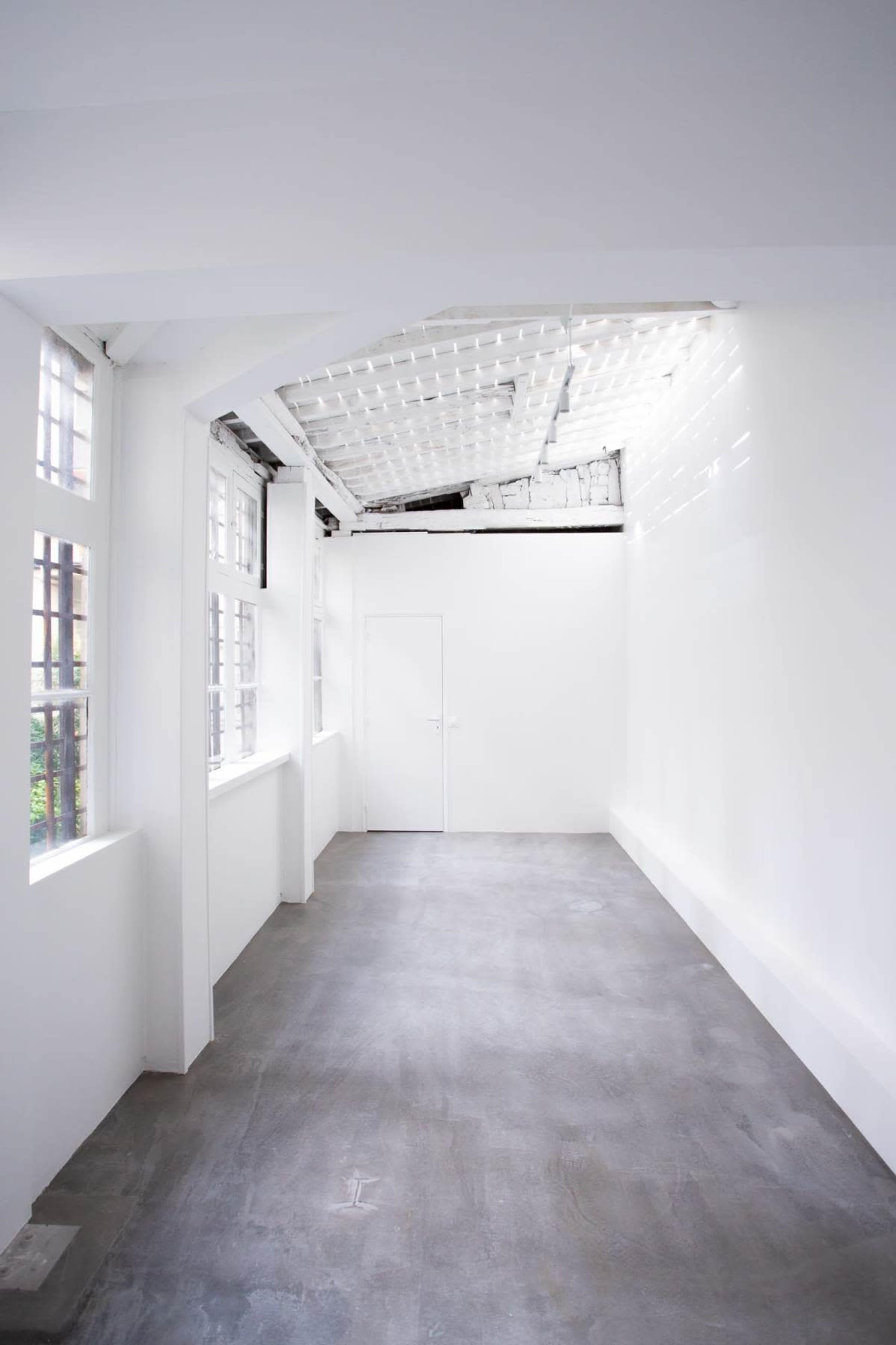 The image shows a minimalist, empty interior room with gray flooring, white walls, and large windows.
