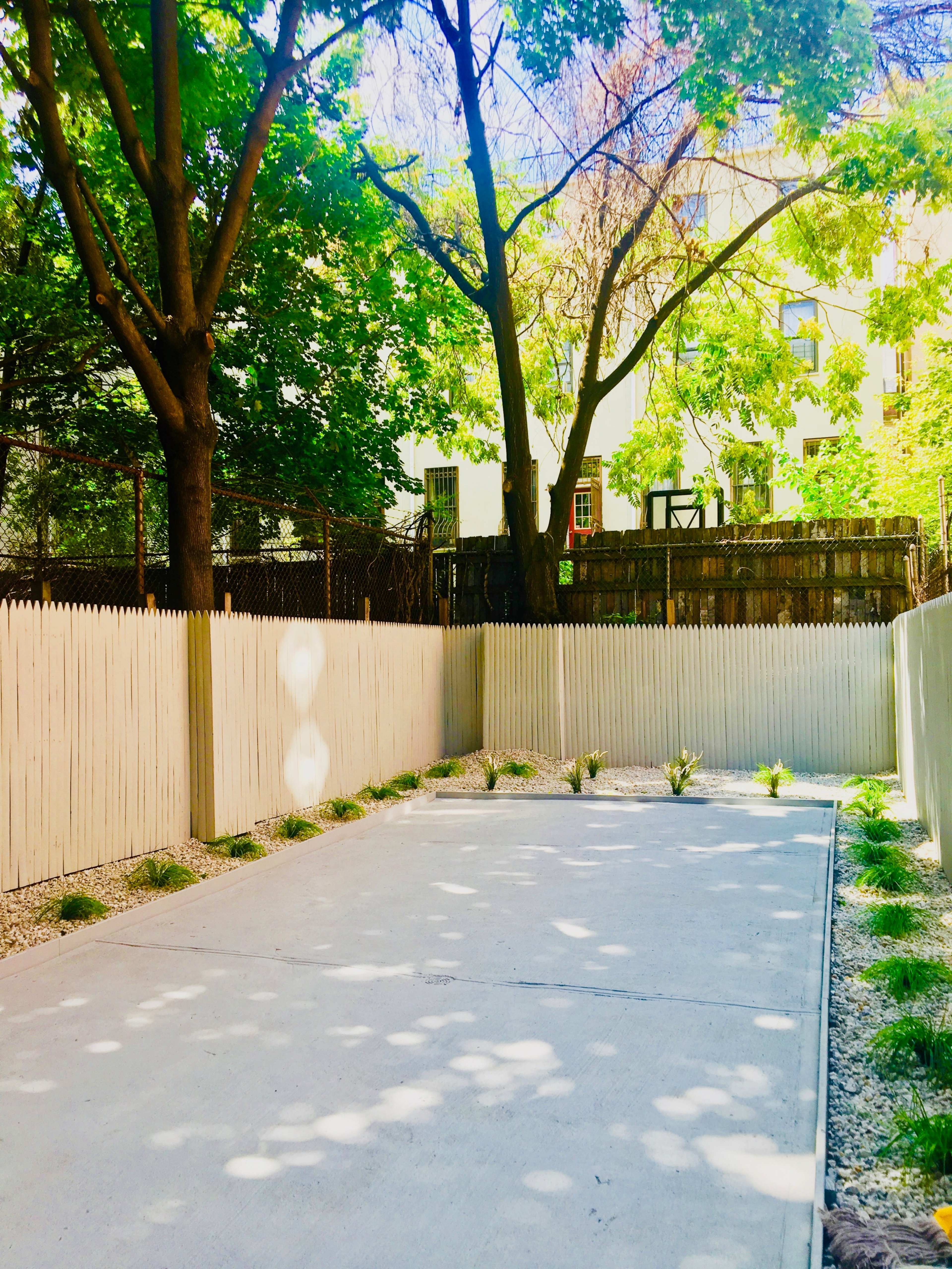 A concrete pathway is bordered by a wooden fence and flanked by greenery in a shaded area.