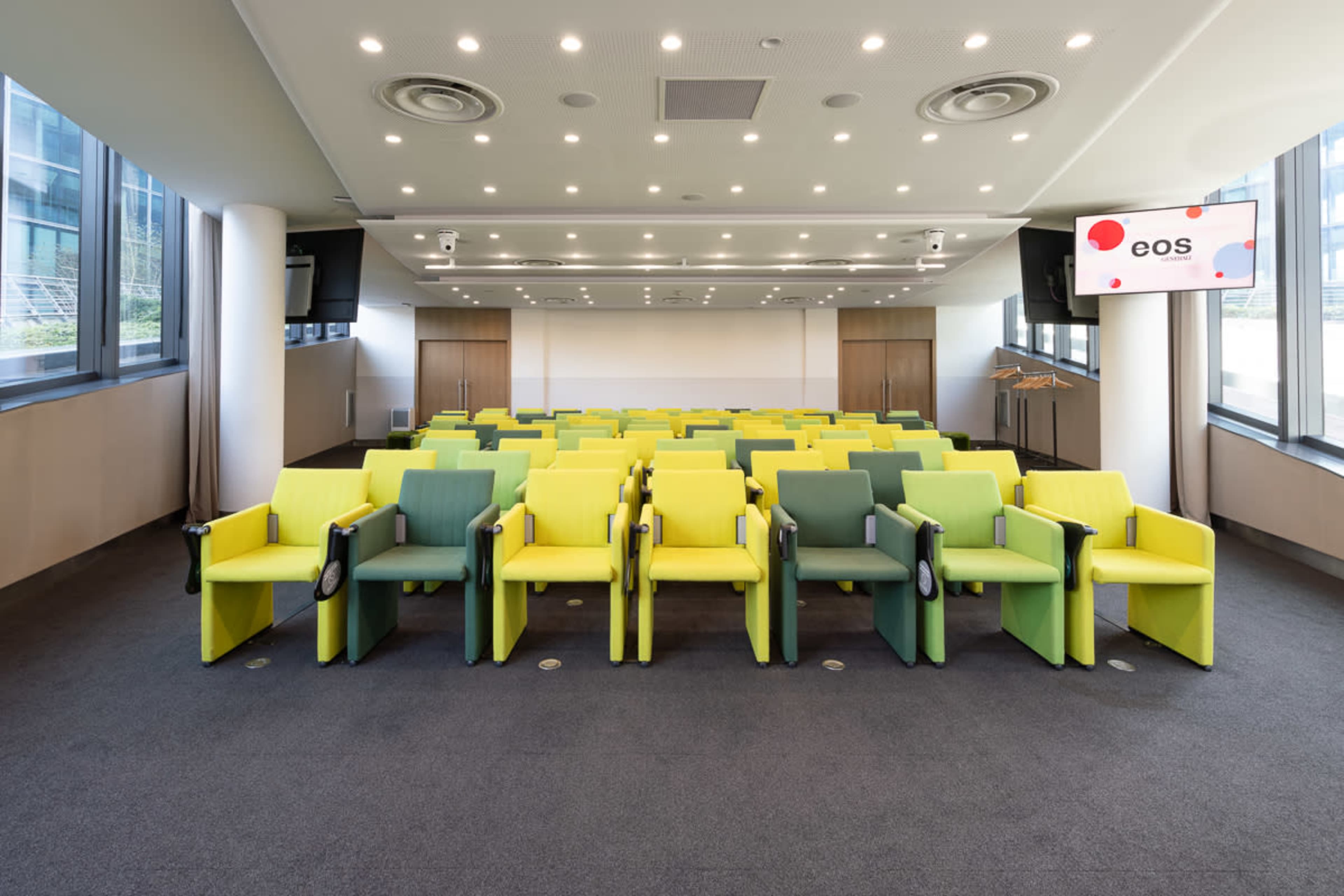 The image shows a conference room with rows of bright green and yellow chairs arranged neatly in front of a screen displaying the word "eos."
