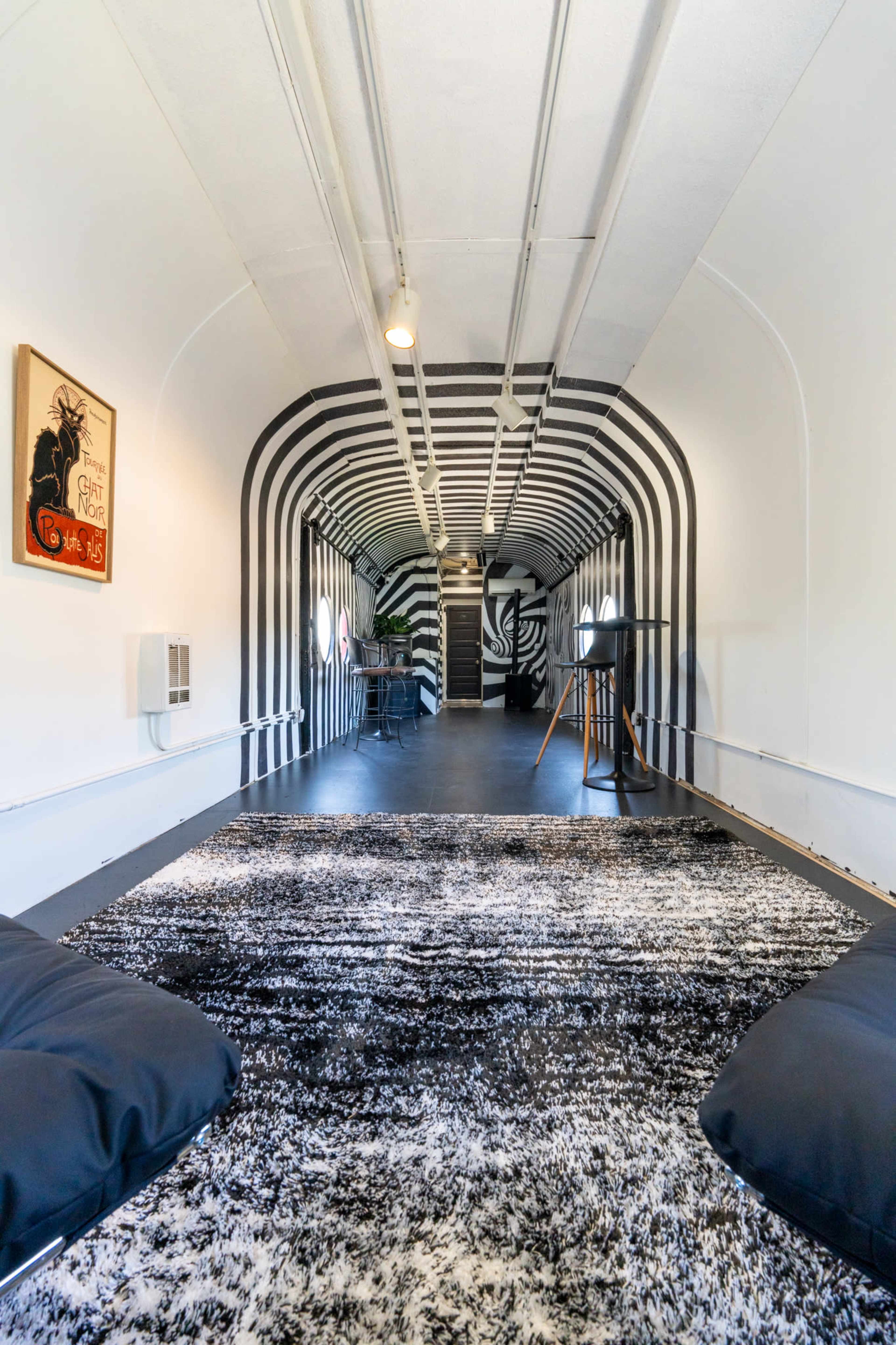 The image shows a narrow corridor with black and white striped walls, leading to a room adorned with minimalist furniture and art.