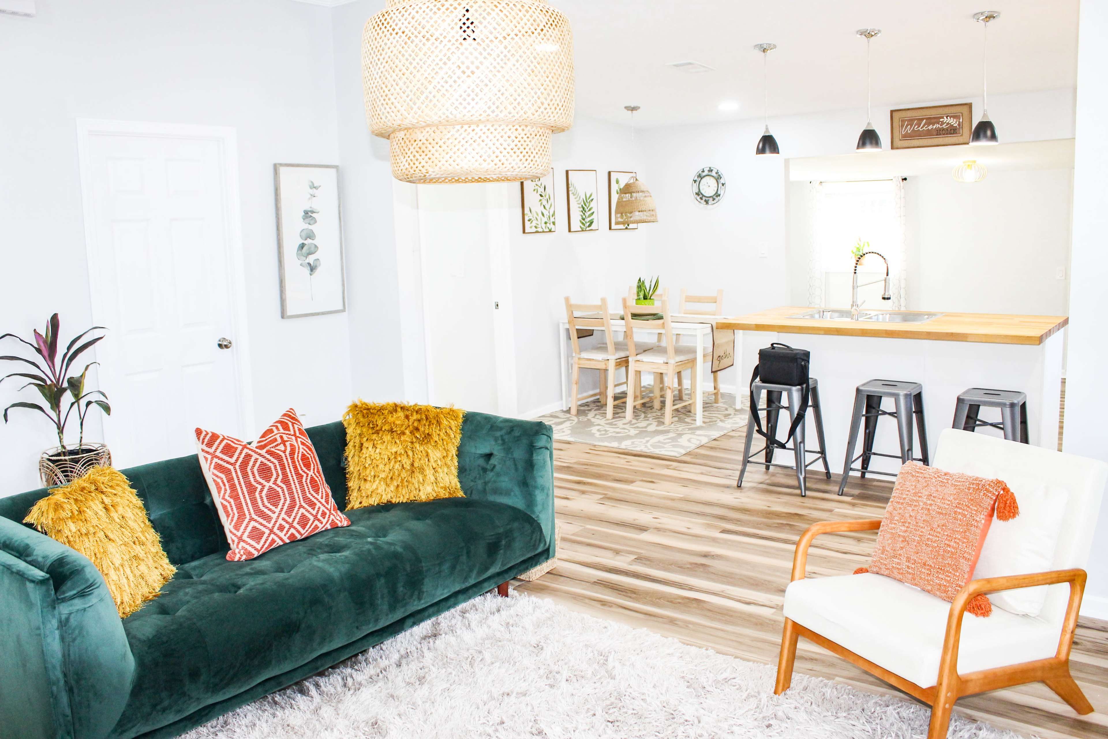 A modern living room with a green couch, decorative pillows, and a white chair, leading to a dining area with a wooden table and stools.