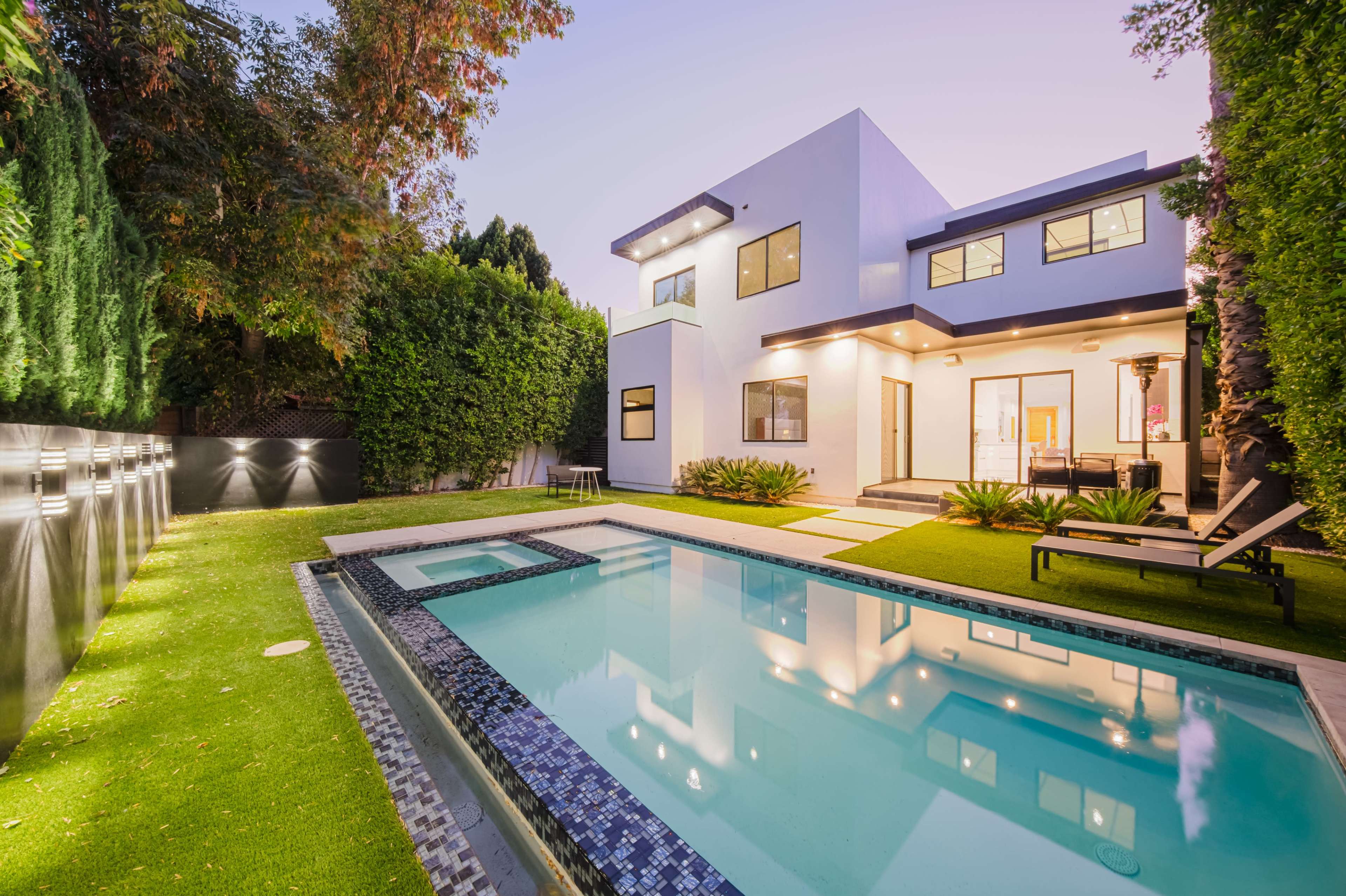 A modern house with large windows is surrounded by greenery and features a swimming pool and a hot tub in the backyard.