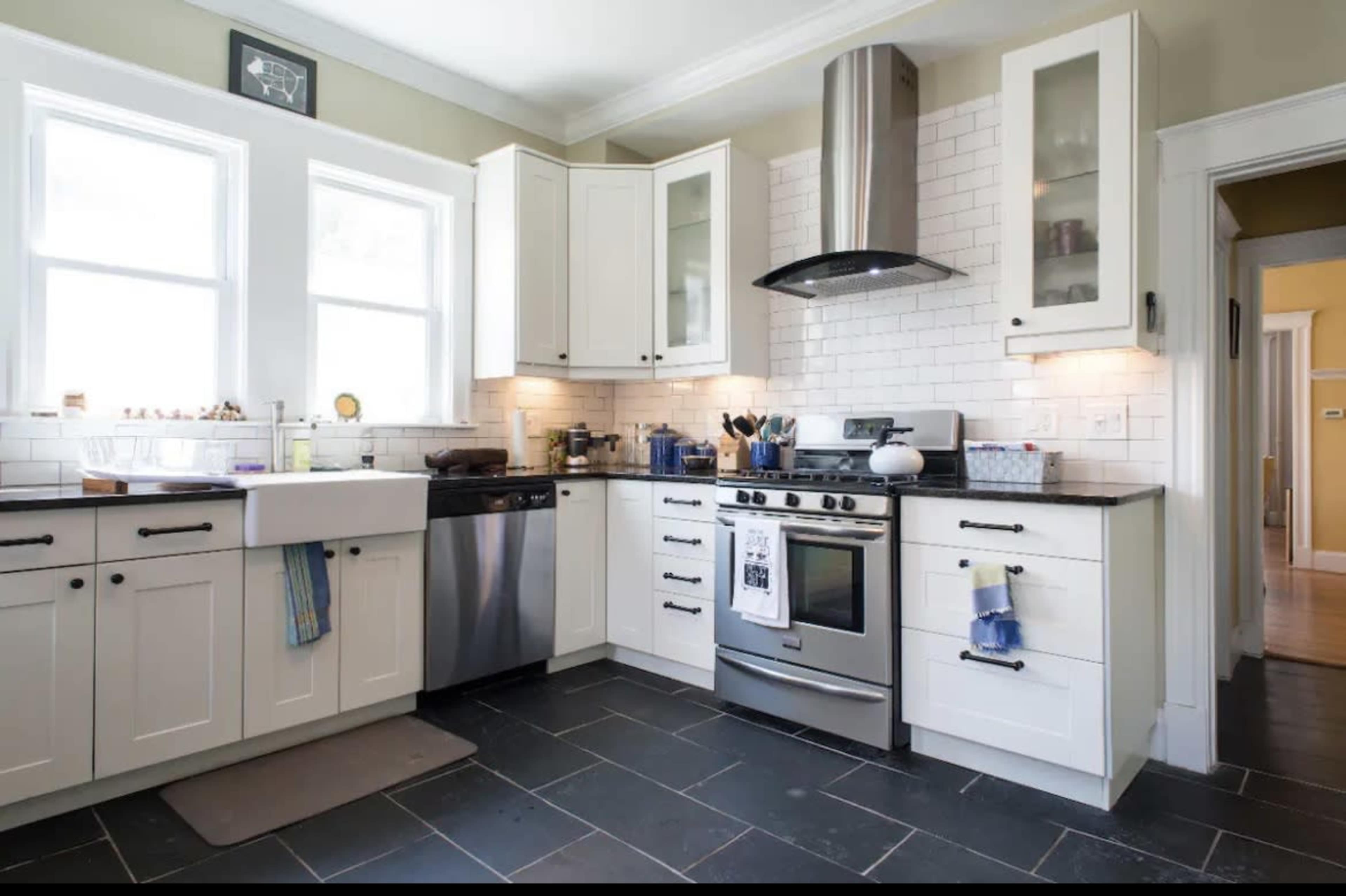 A modern kitchen features white cabinetry, stainless steel appliances, and a black countertop, with a double sink under large windows.