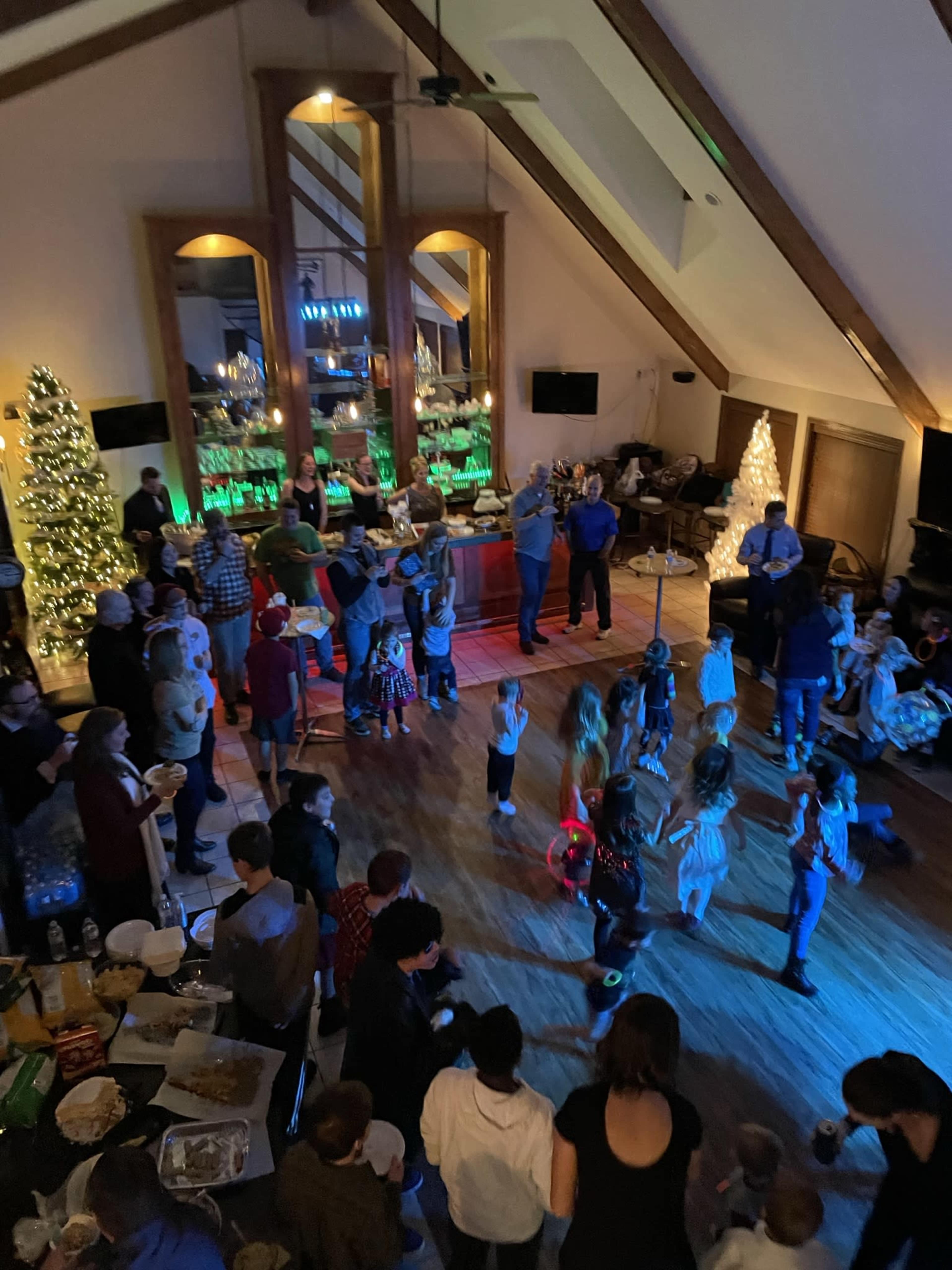 A large group of people, including children and adults, are dancing and socializing in a festive indoor space decorated with Christmas trees and lighting.