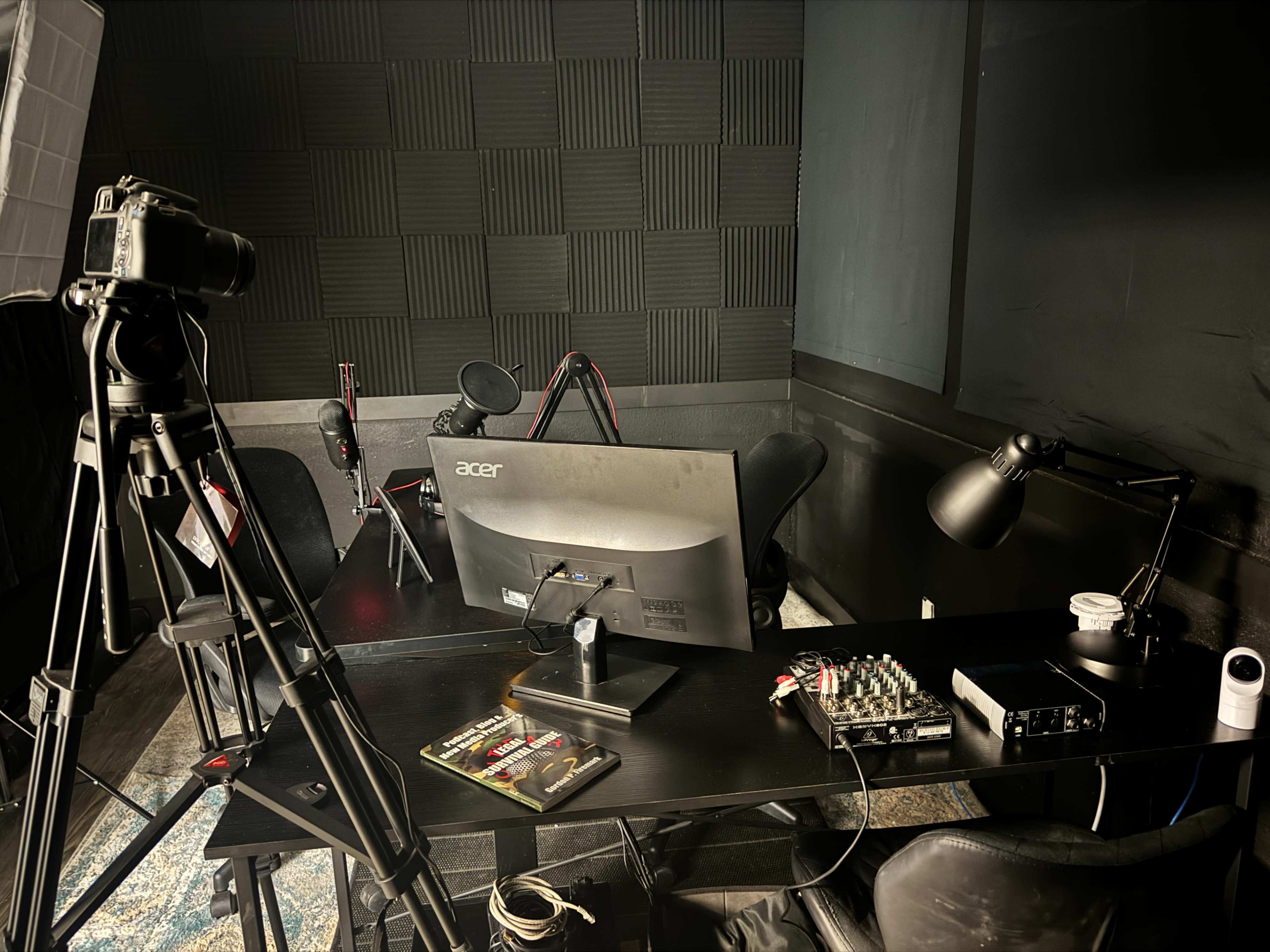 The image shows a recording studio setup featuring a camera on a tripod, a computer monitor, mixing equipment, and a few chairs arranged in a dimly lit space with soundproofing panels on the walls.