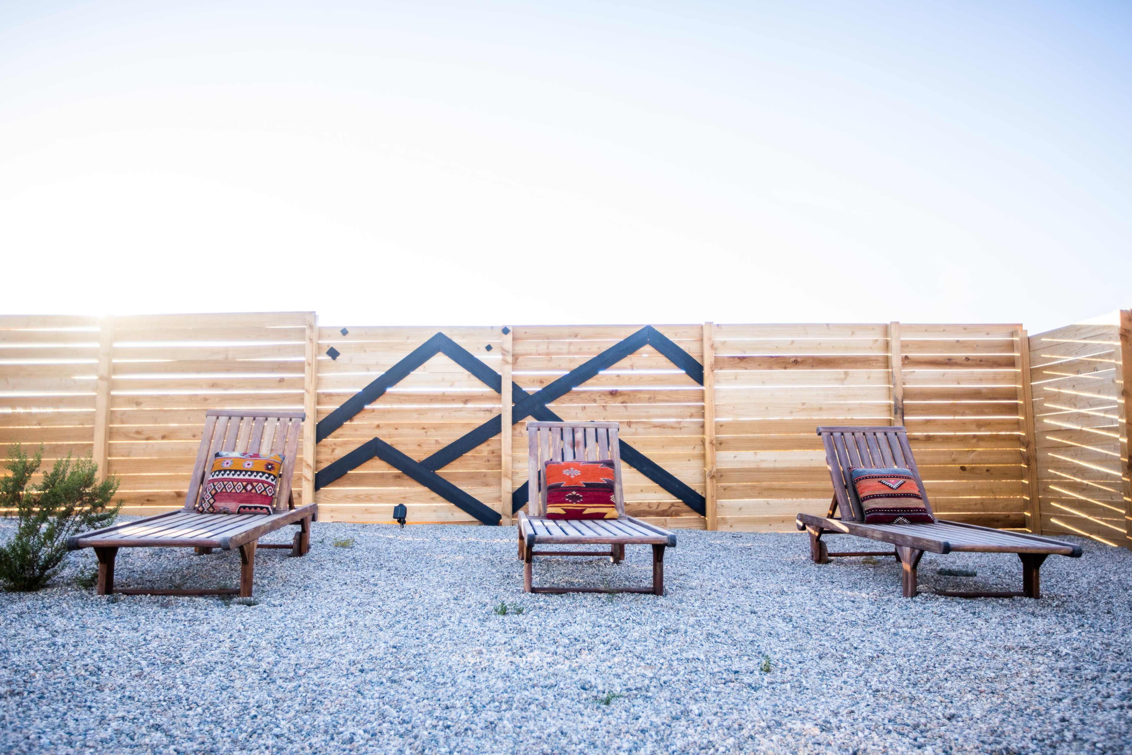Three wooden lounge chairs with patterned cushions sit on a gravel patio in front of a wooden fence featuring a geometric design.