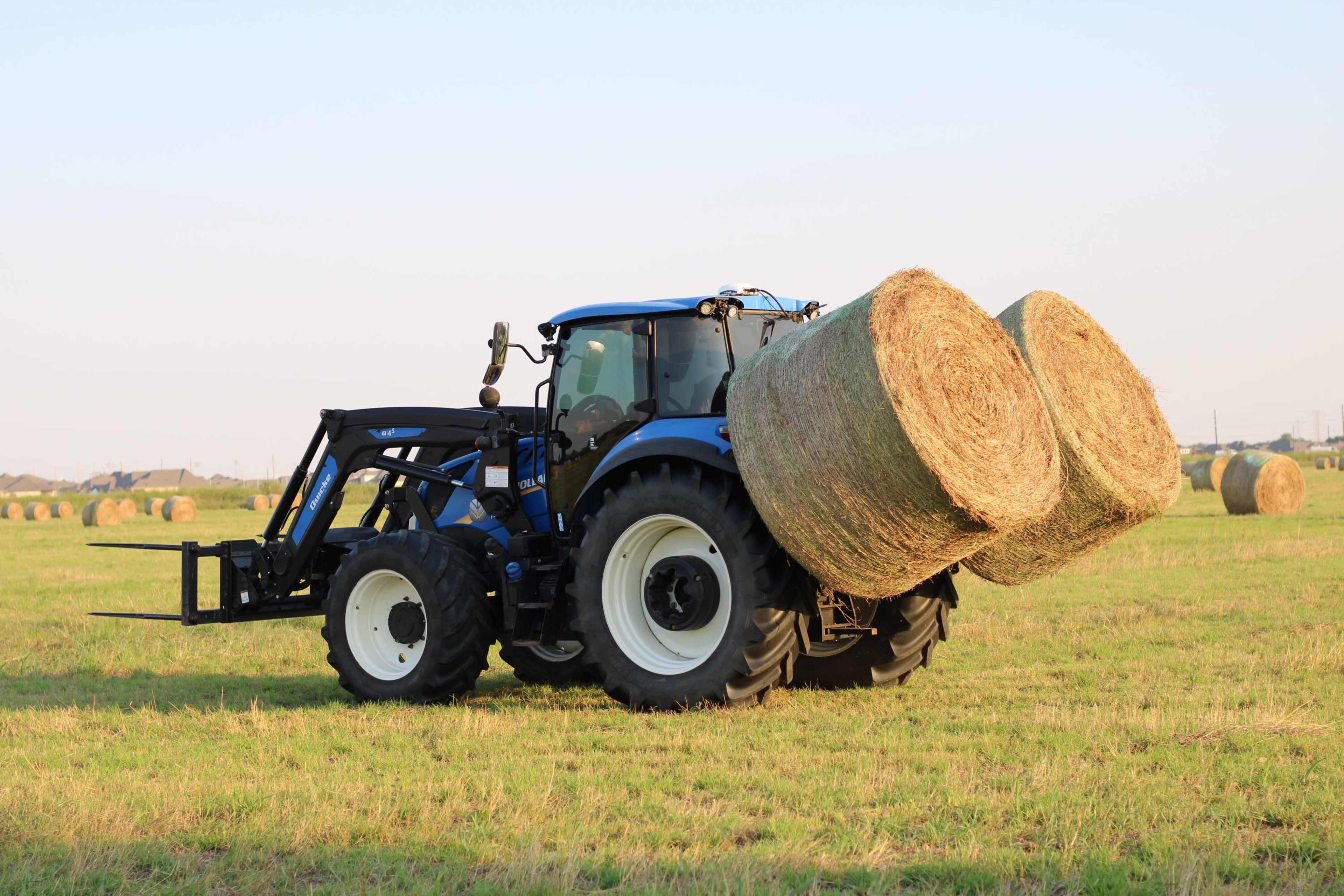 A blue tractor carries two large round hay bales across a grassy field.