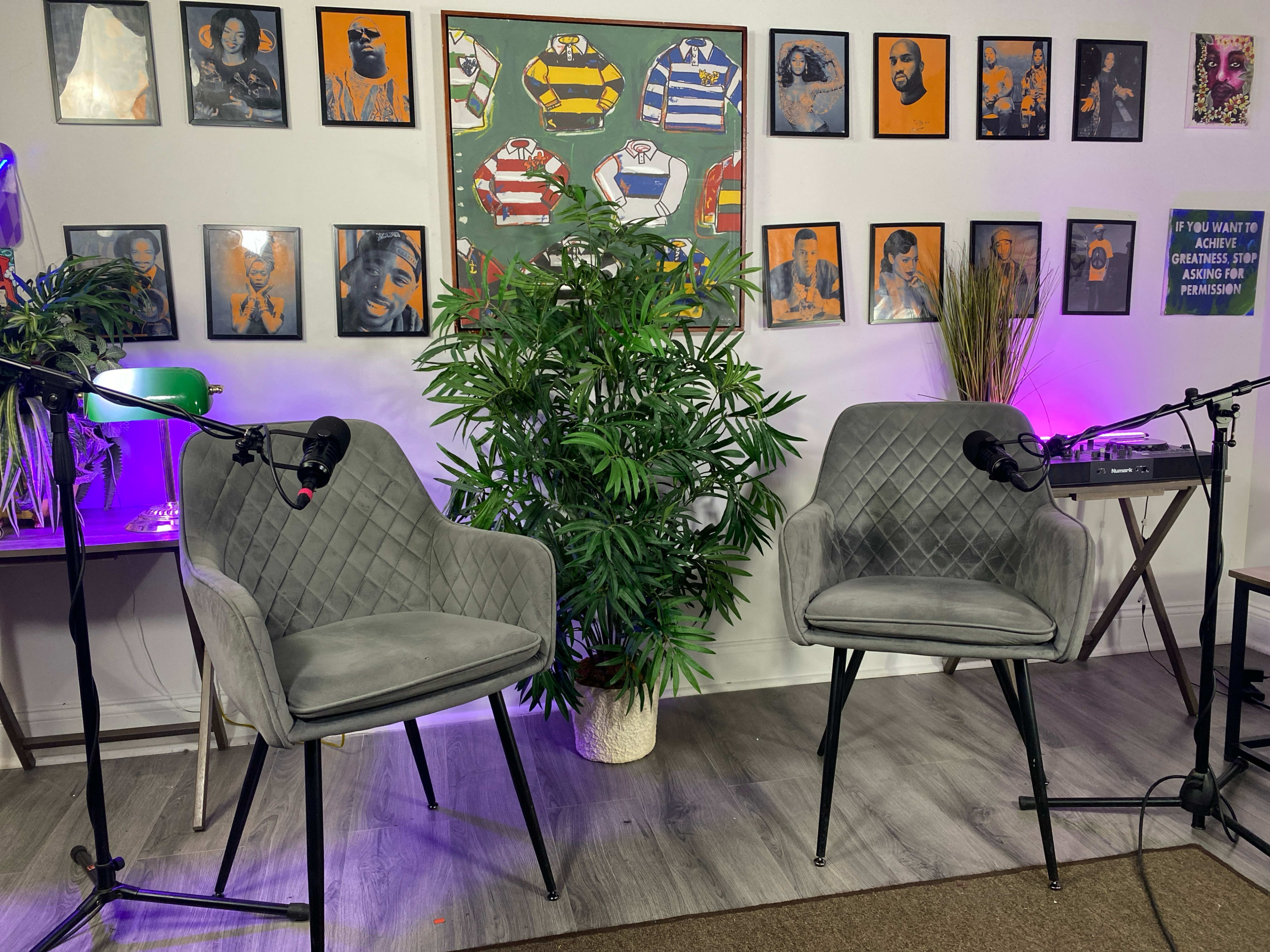 The image shows a podcast recording setup with two empty gray chairs facing each other, microphones attached, and a wall adorned with framed art and sports jerseys.