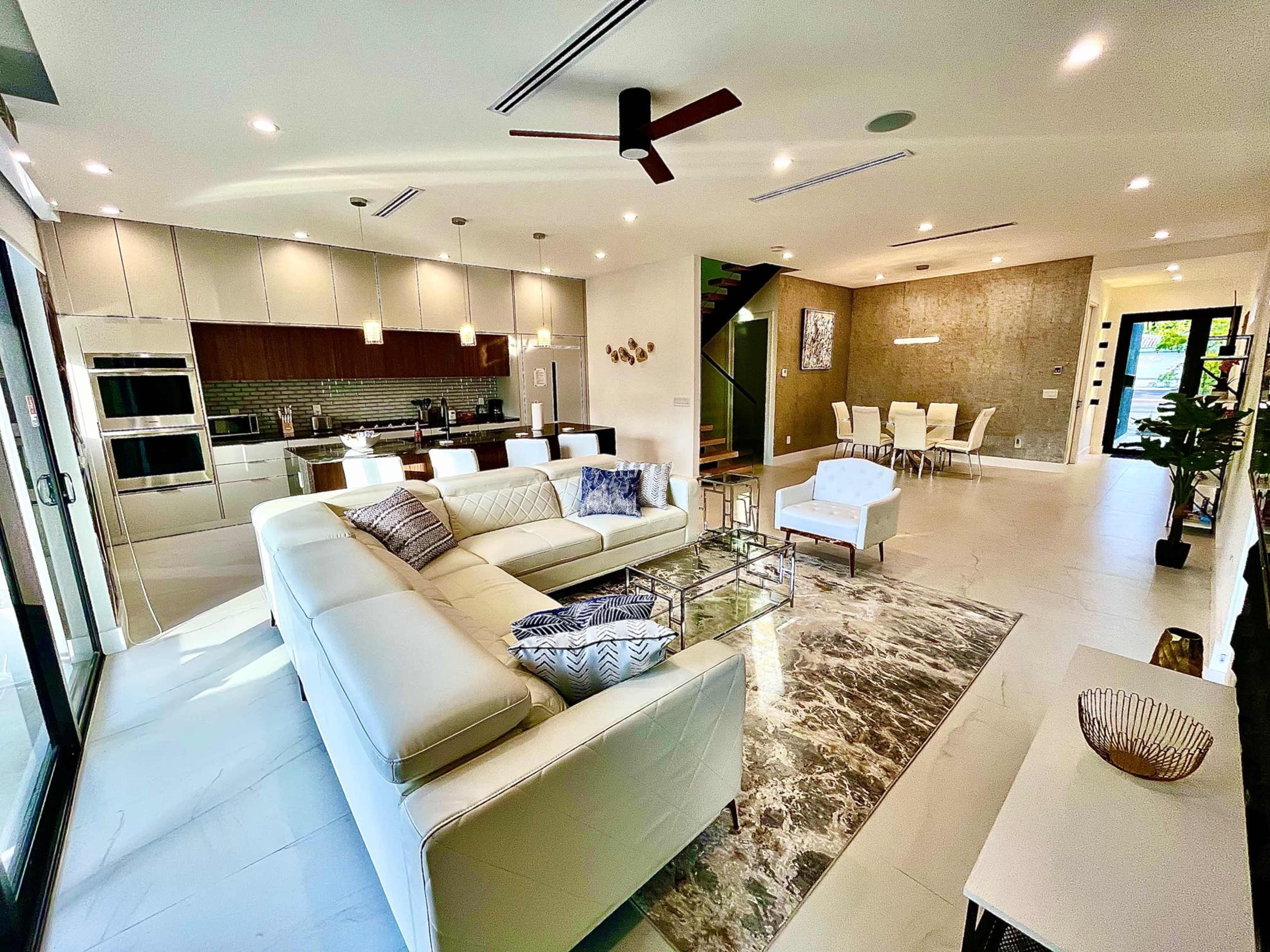 The image shows a modern living room connected to a kitchen and dining area, featuring sleek furniture, large windows, and a marble floor.