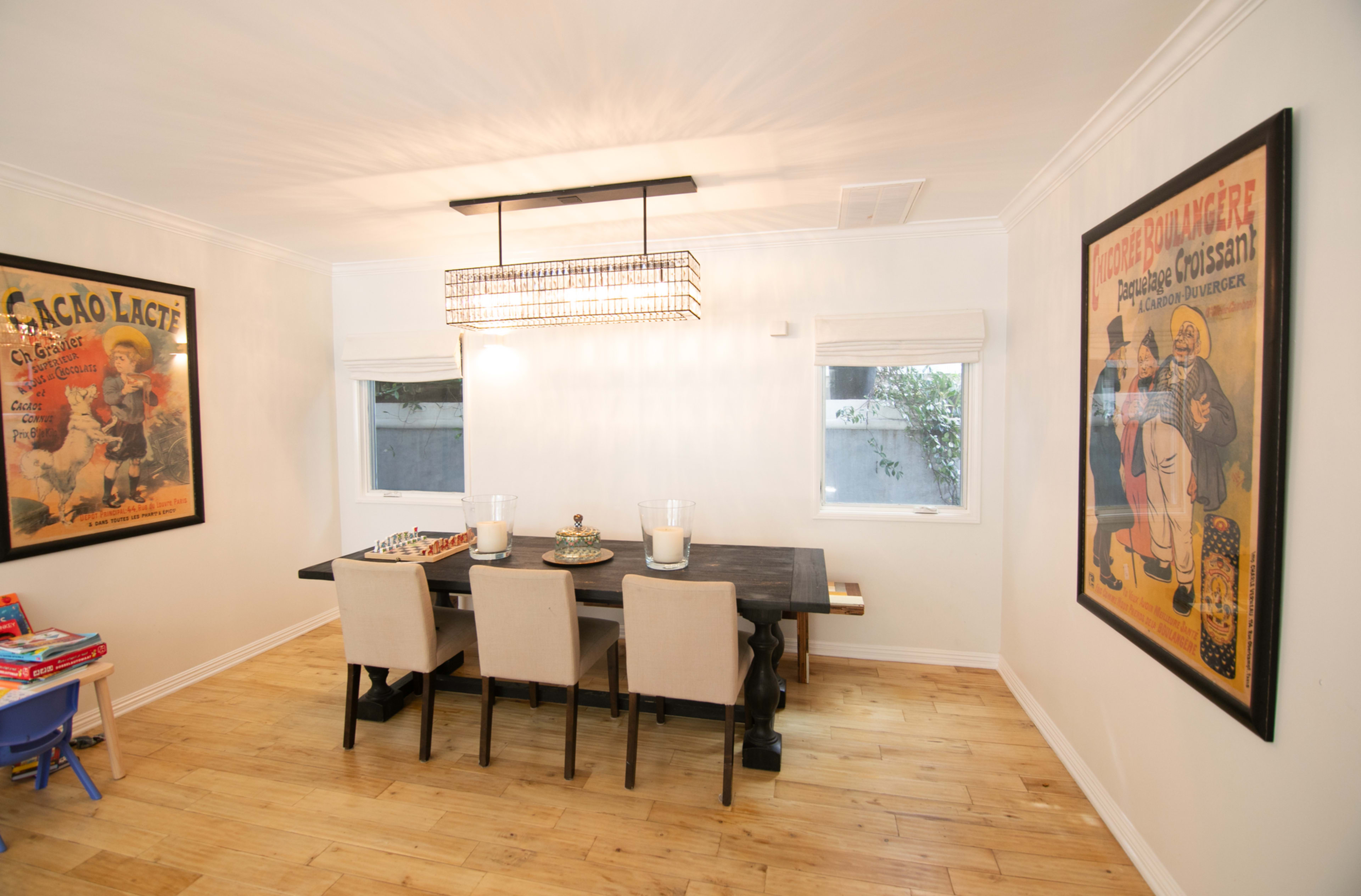 A dining room features a central table surrounded by four chairs, with artwork on the walls and large windows allowing natural light.