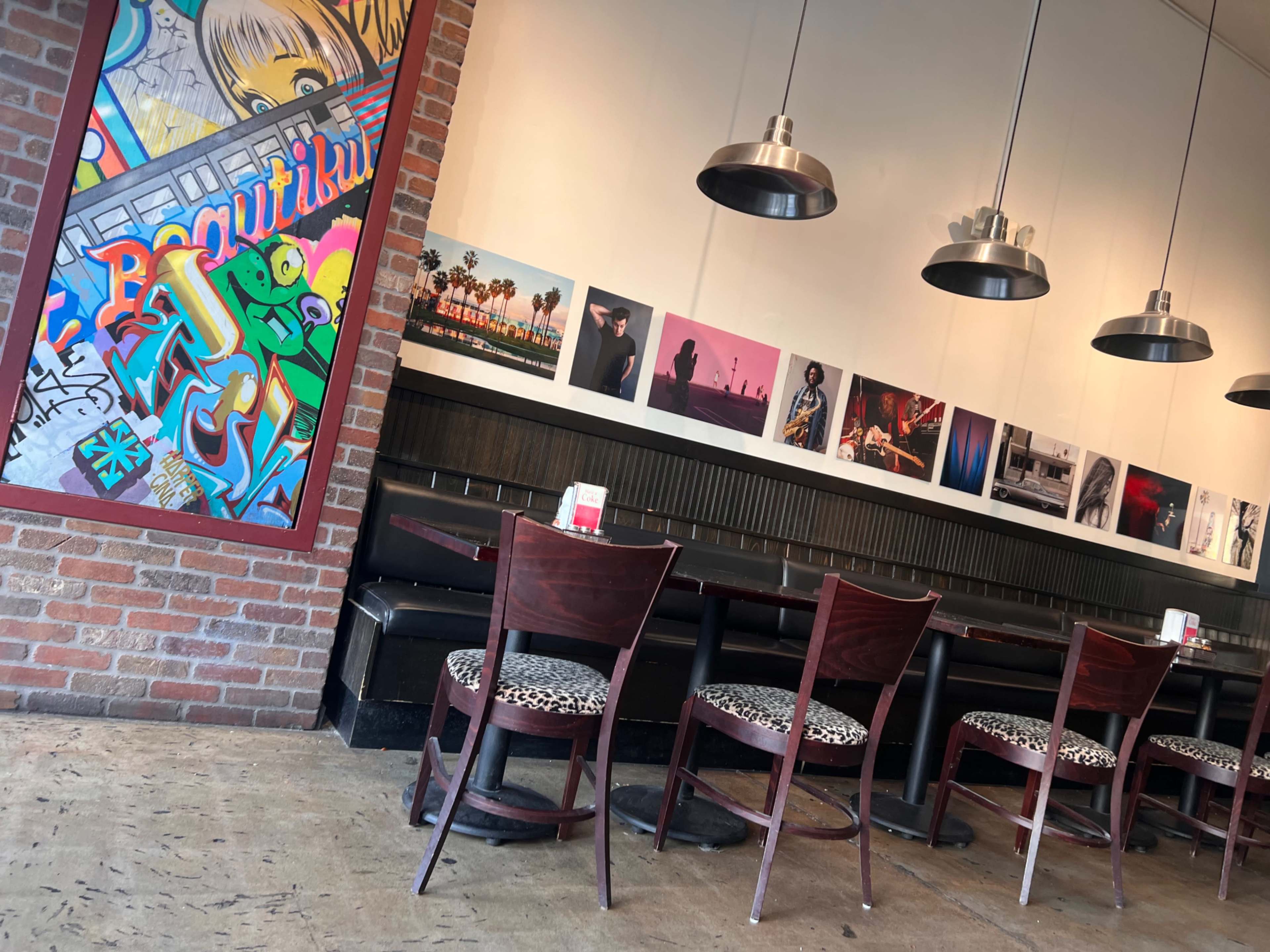 The image shows a restaurant interior featuring a colorful mural, several framed photographs on the wall, and wooden tables with chairs arranged along a brick wall.