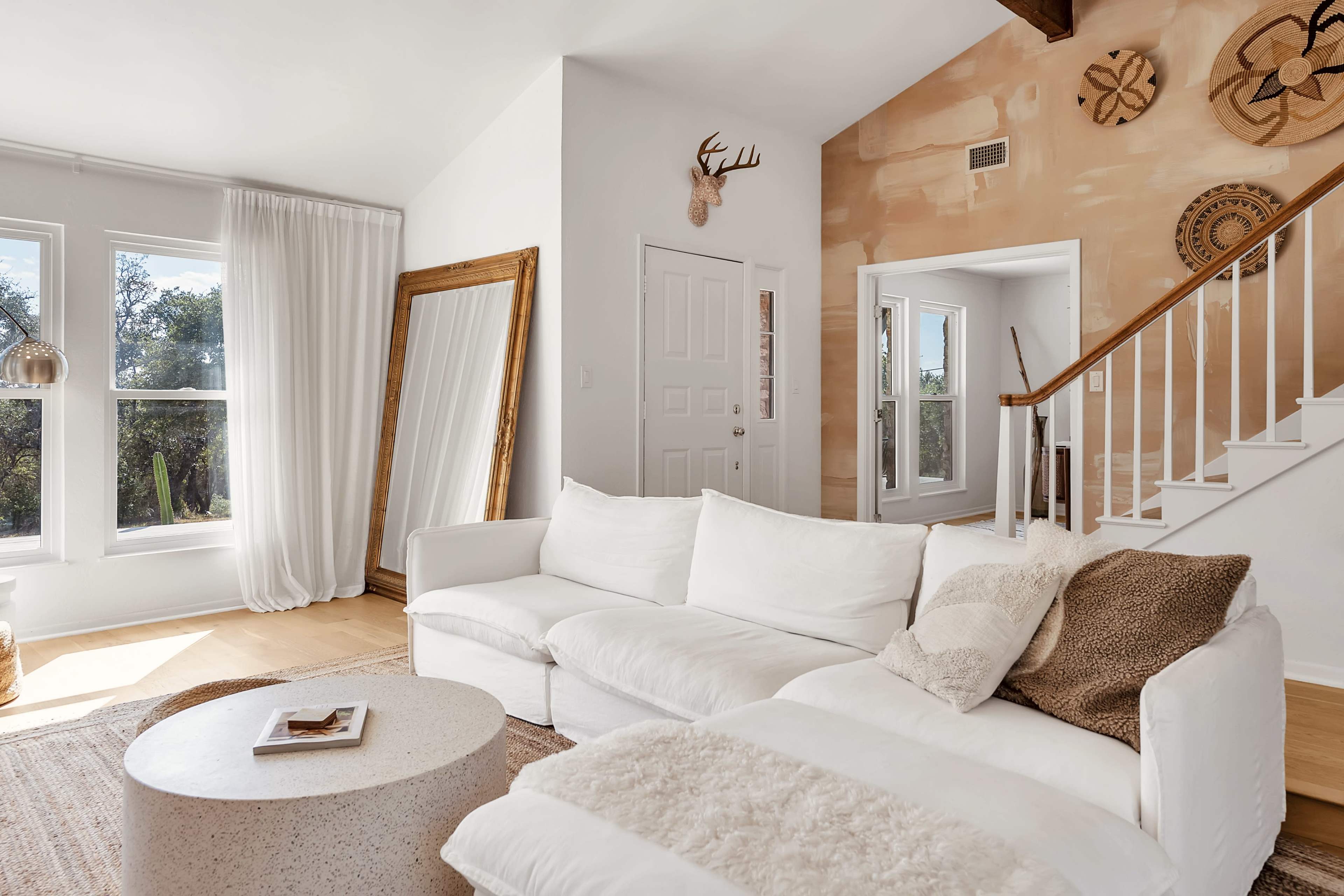 The living room features a white sectional sofa, a round coffee table, large windows, and a staircase leading to another area.