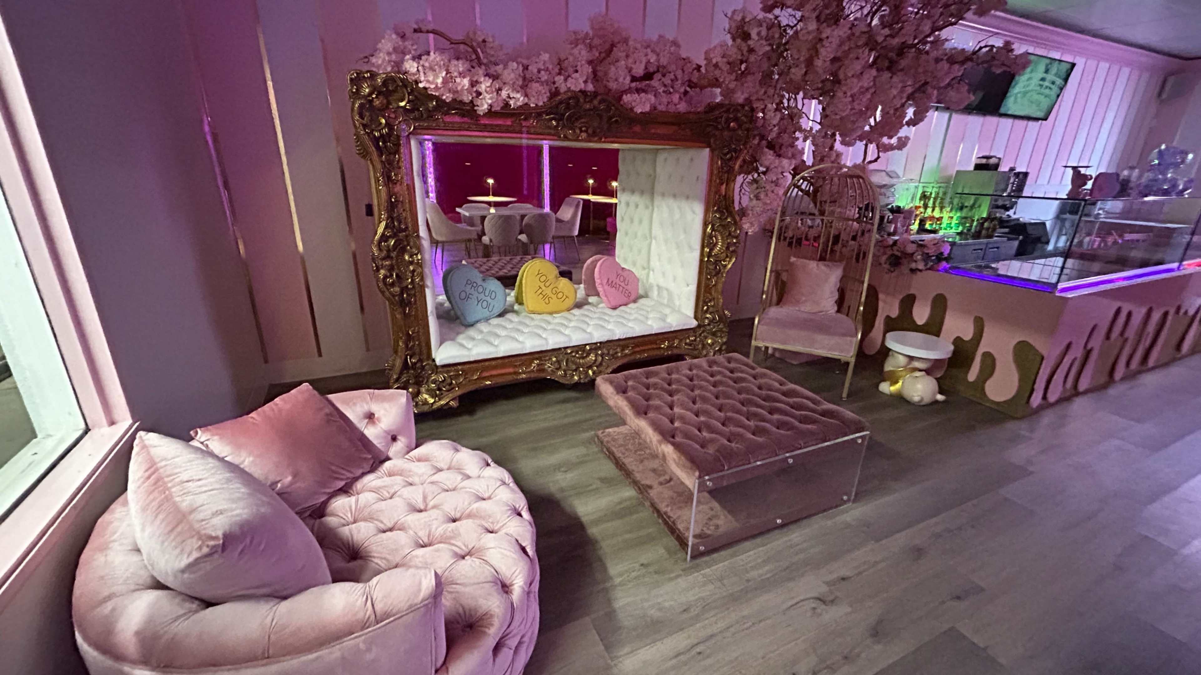 The image shows a cozy lounge area featuring plush pink seating, a decorative mirror surrounded by artificial cherry blossoms, and a small table in the background.