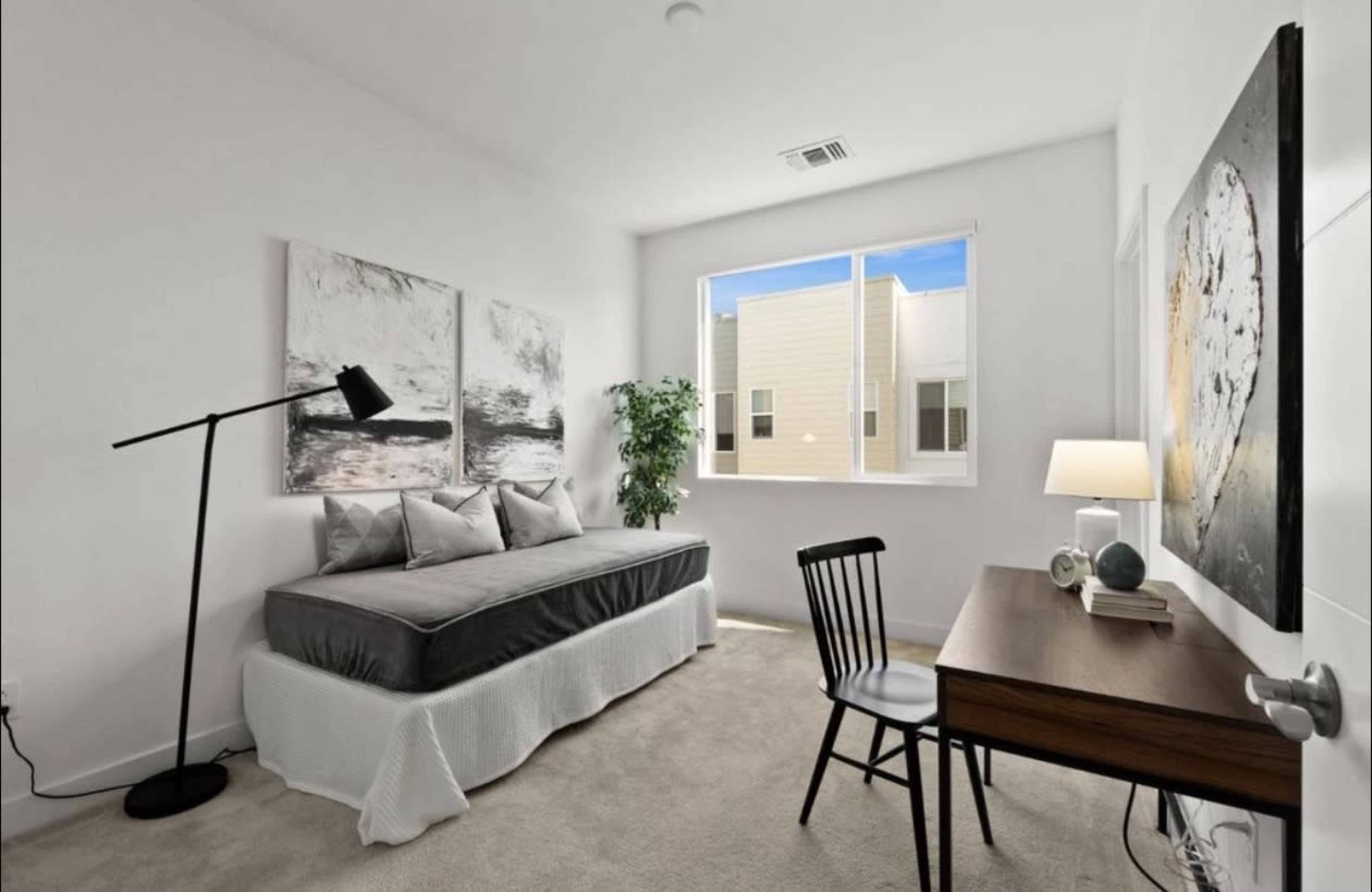 The image shows a minimalist bedroom featuring a bed with a gray mattress and bedding, a black chair, a desk, and two decorative wall artworks.