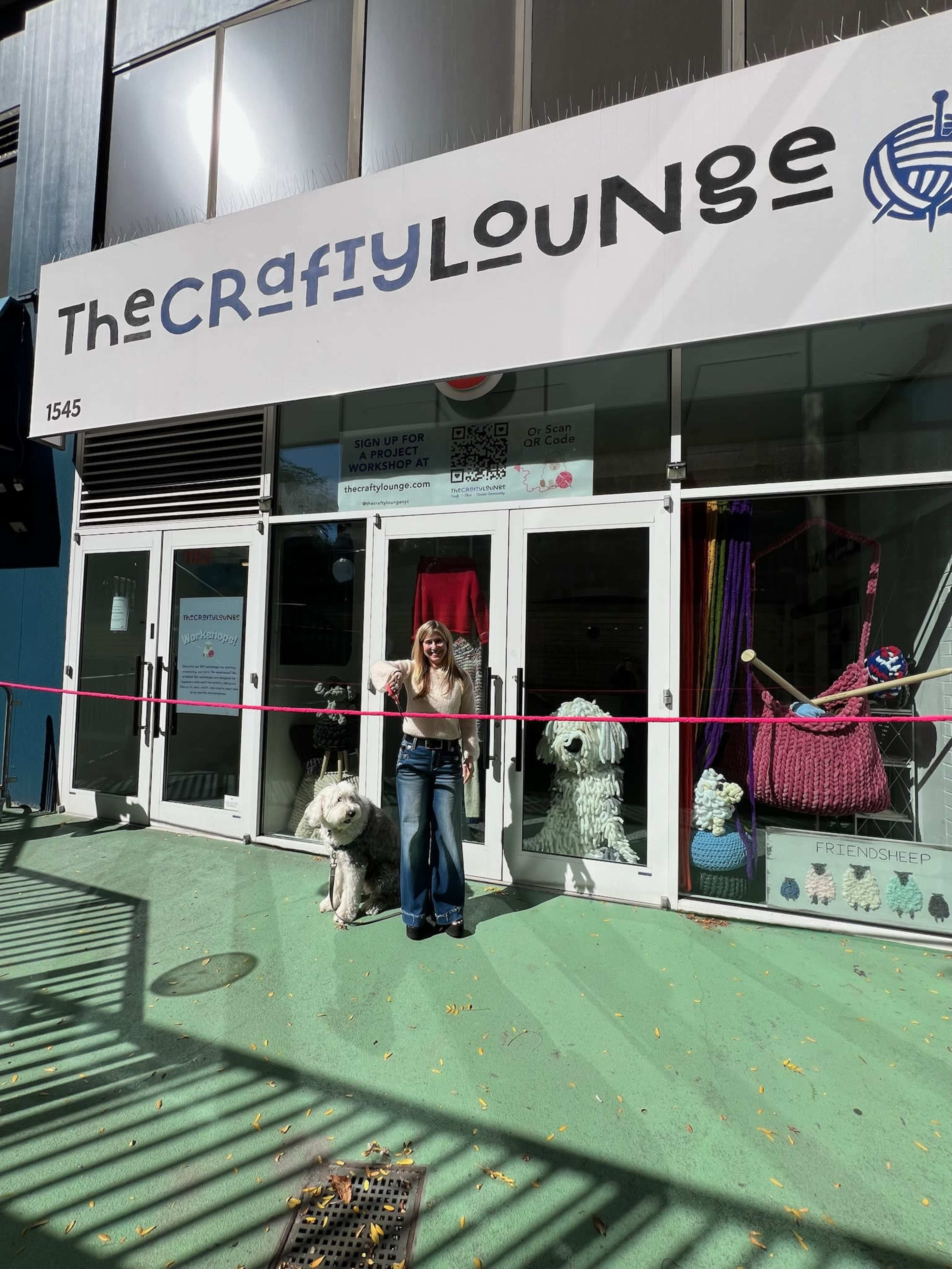 A woman stands with a large dog in front of a crafting store named "The Crafty Lounge" with a colorful display and a ribbon across the entrance.