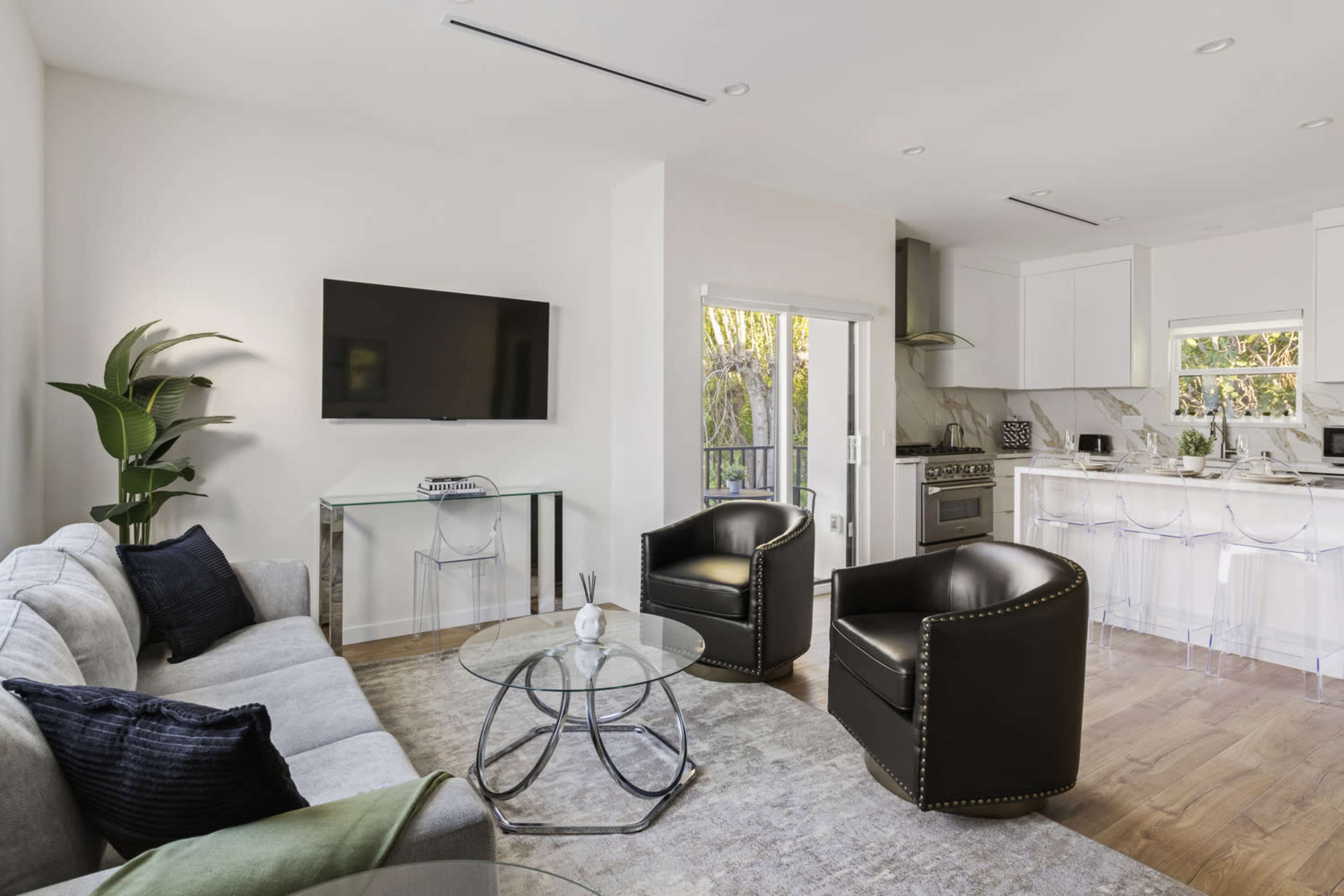 The image shows a modern living area with a gray sofa, two black chairs, a glass coffee table, a wall-mounted TV, and a kitchen visible in the background.