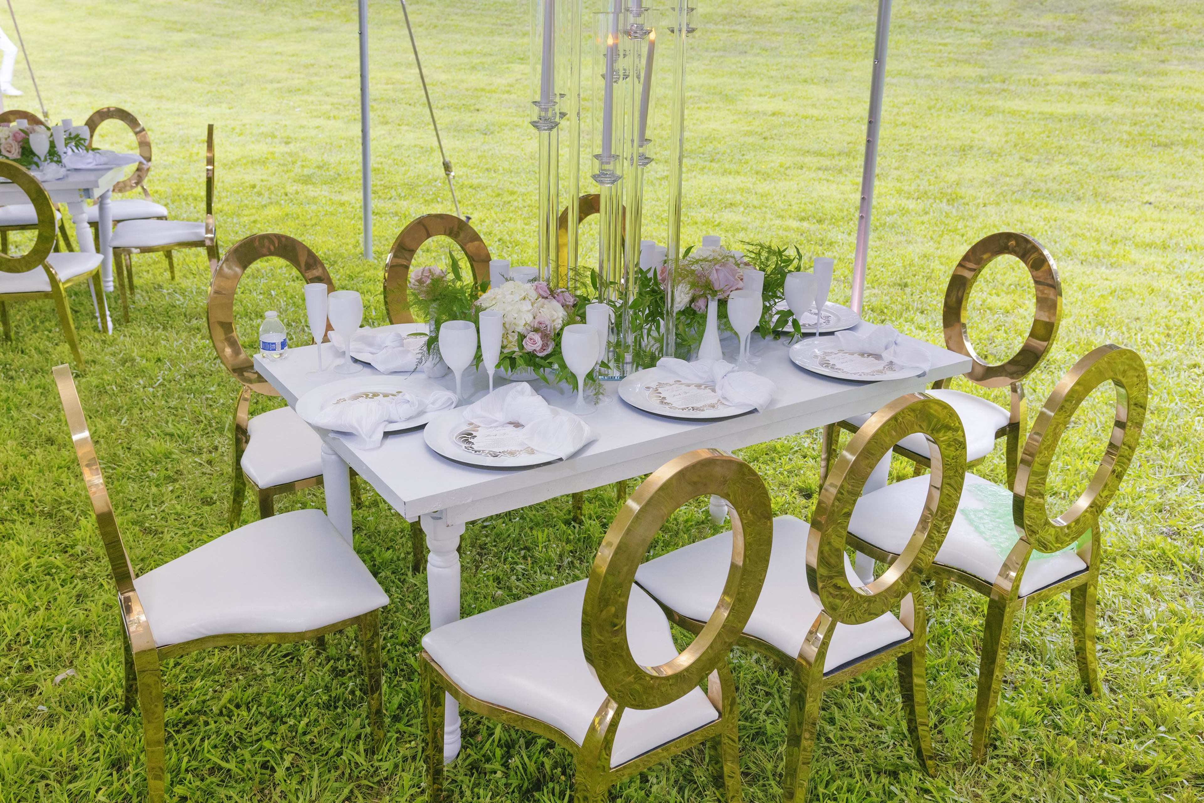 A white table is set with plates and floral arrangements, surrounded by gold-accented chairs on a grassy area.