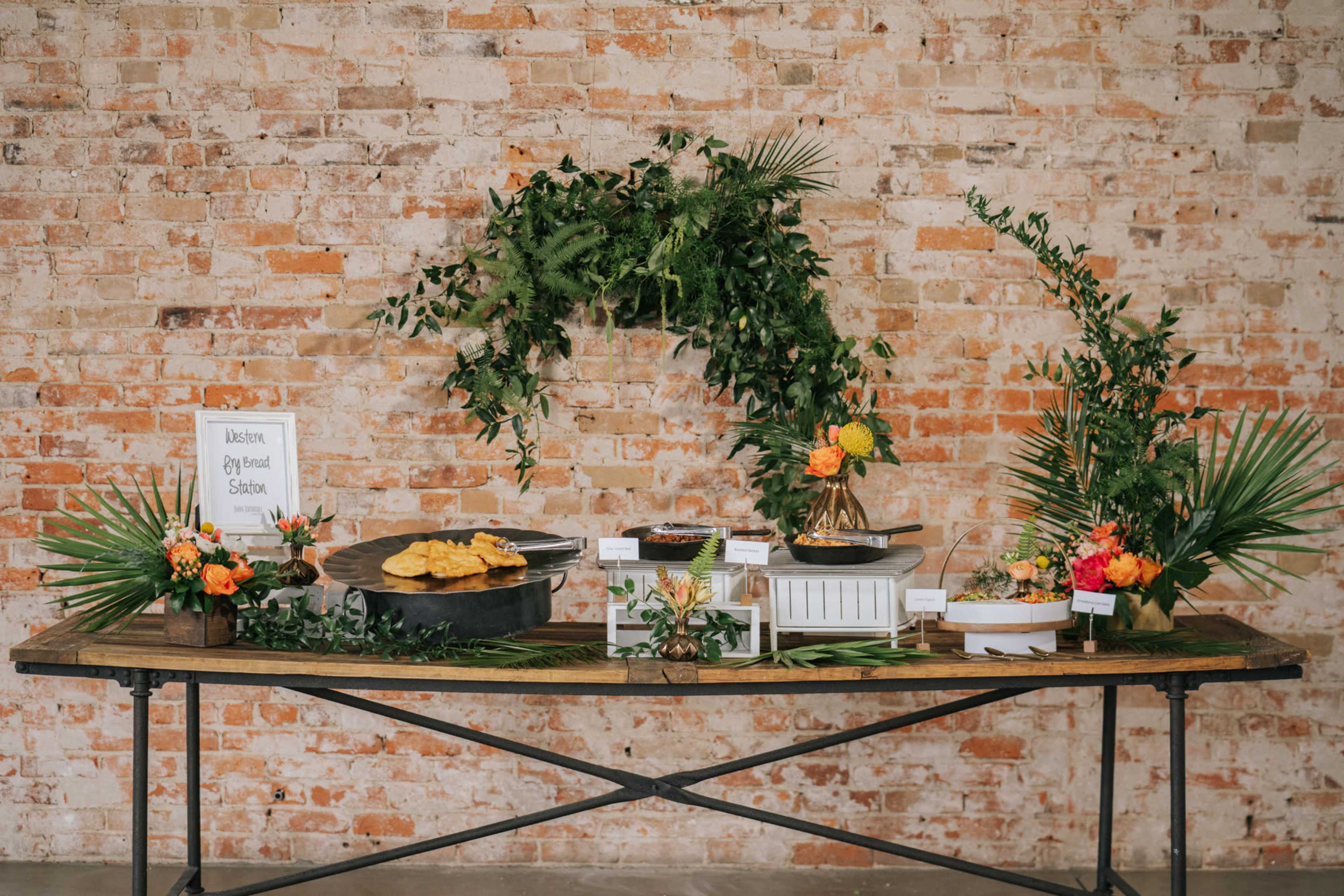 A buffet table adorned with various platters of food and floral decorations is set against a rustic brick wall.