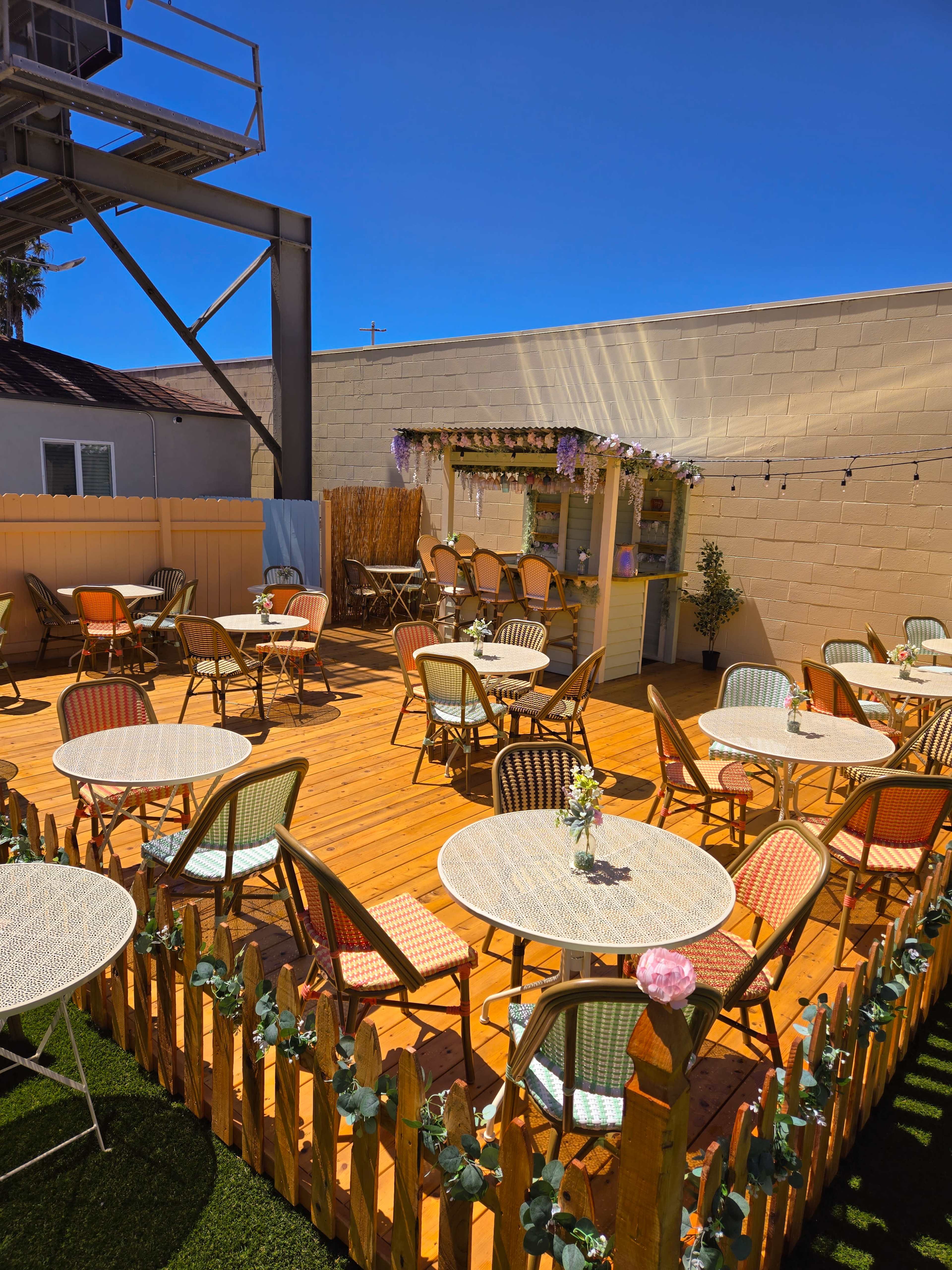 A wooden patio area with several round tables and chairs, surrounded by a low fence and decorated with string lights and flowers.