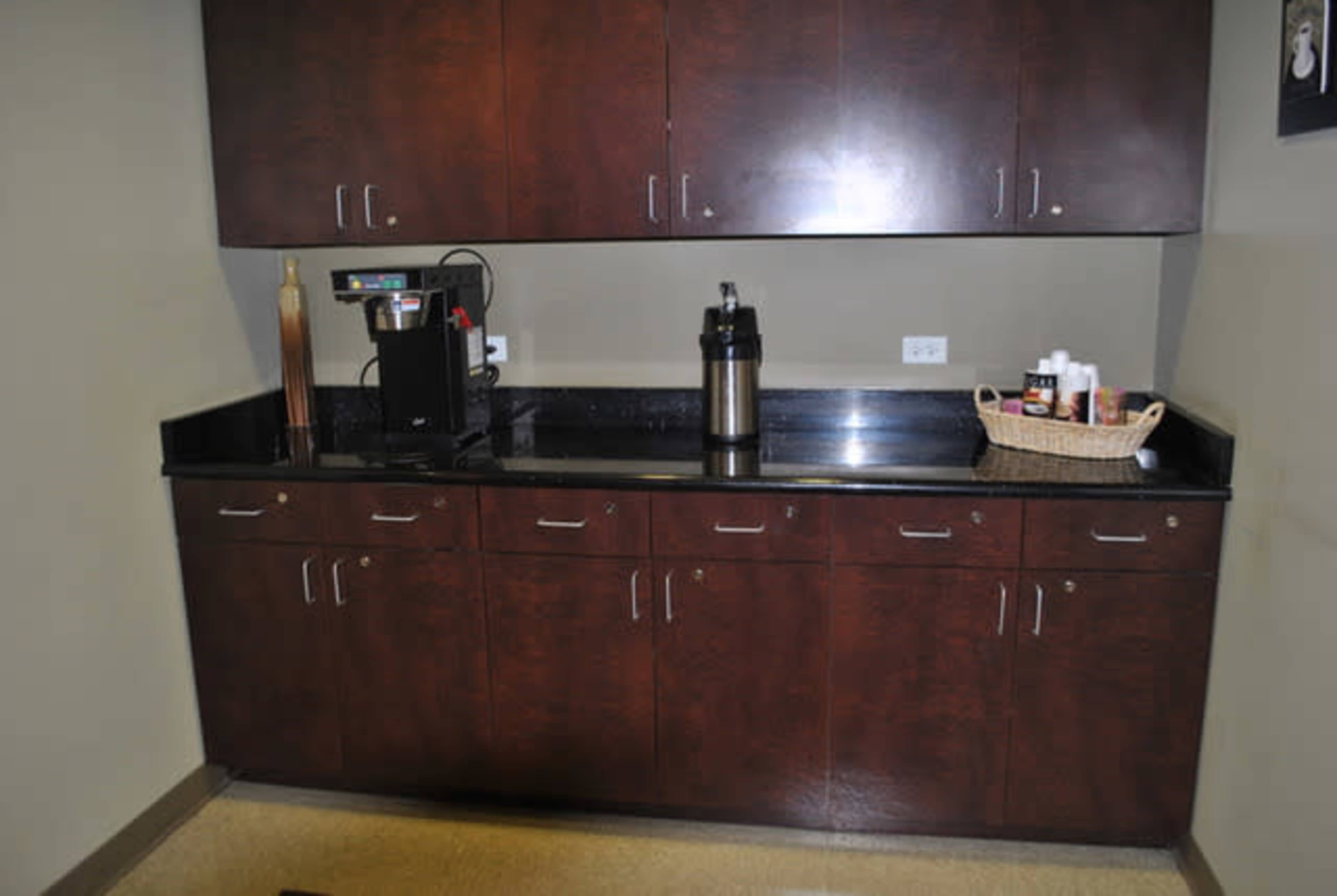 A kitchen area with dark wood cabinets above a black countertop, featuring a coffee machine and a thermos next to a basket of condiments.