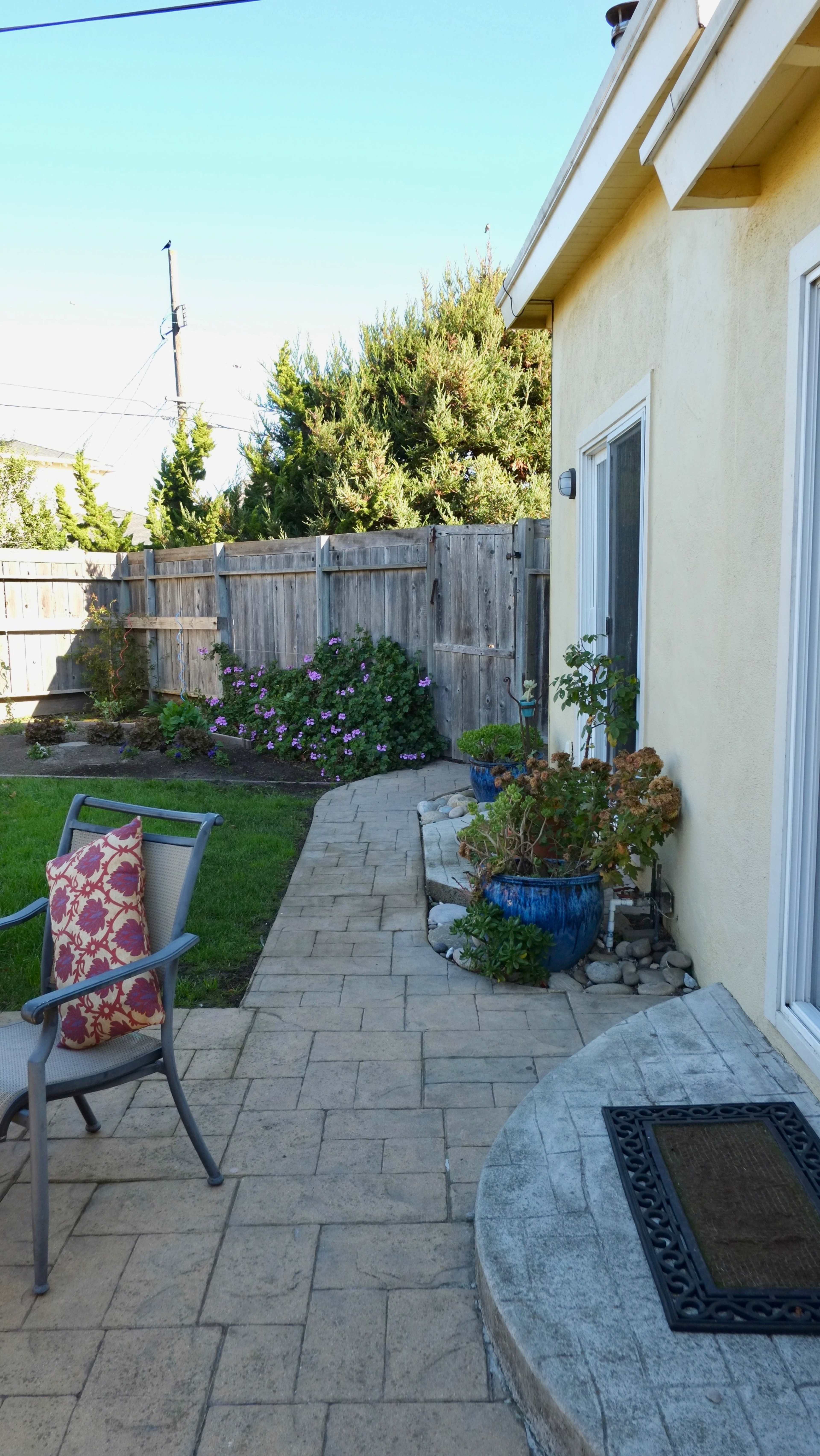 The image shows a paved walkway leading to the back of a yellow house, bordered by green grass and flower beds.