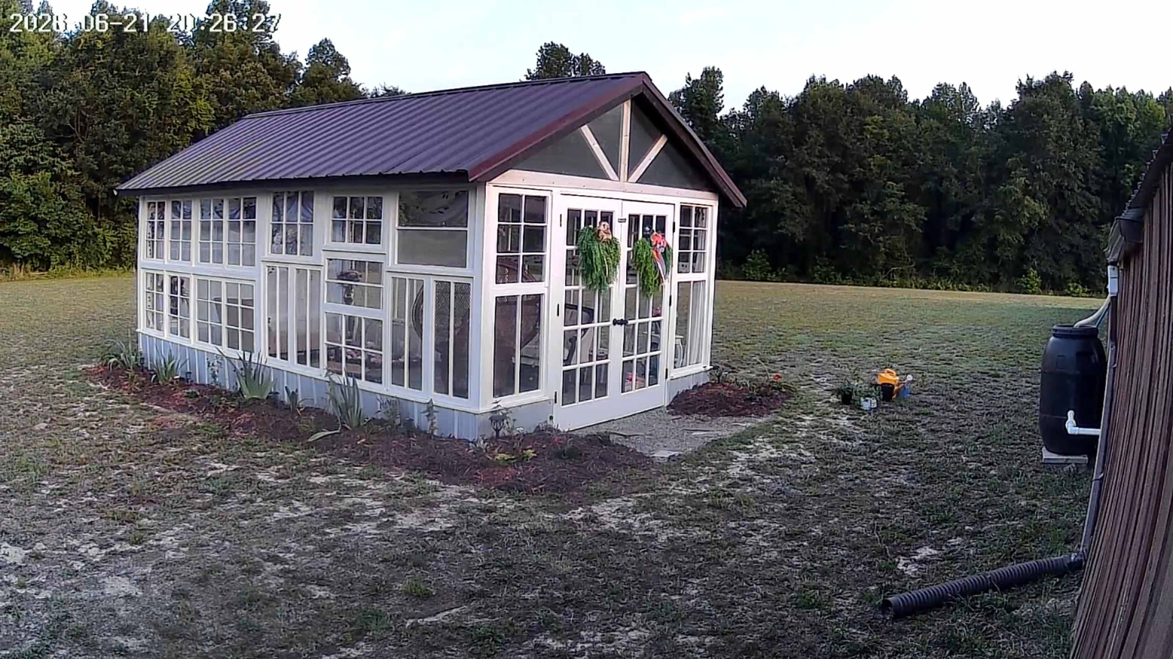 The image shows a glass greenhouse with a metal roof situated in a grassy area, surrounded by trees in the background.